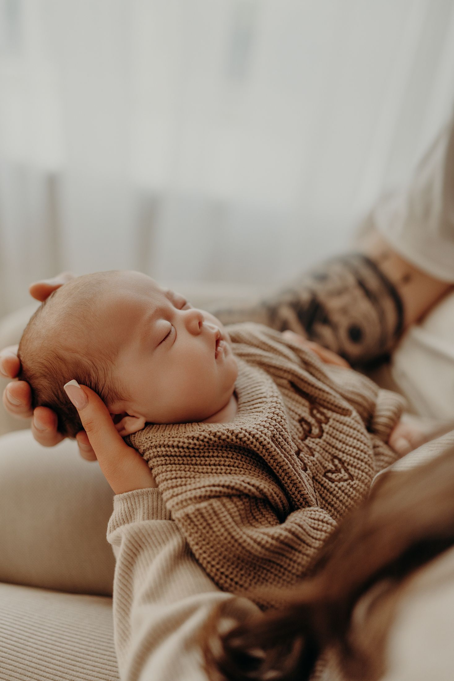 A sleeping newborn baby wearing a cozy beige knit sweater from Grays.bespoke Love is gently cradled in parents hands, newborn session dublin