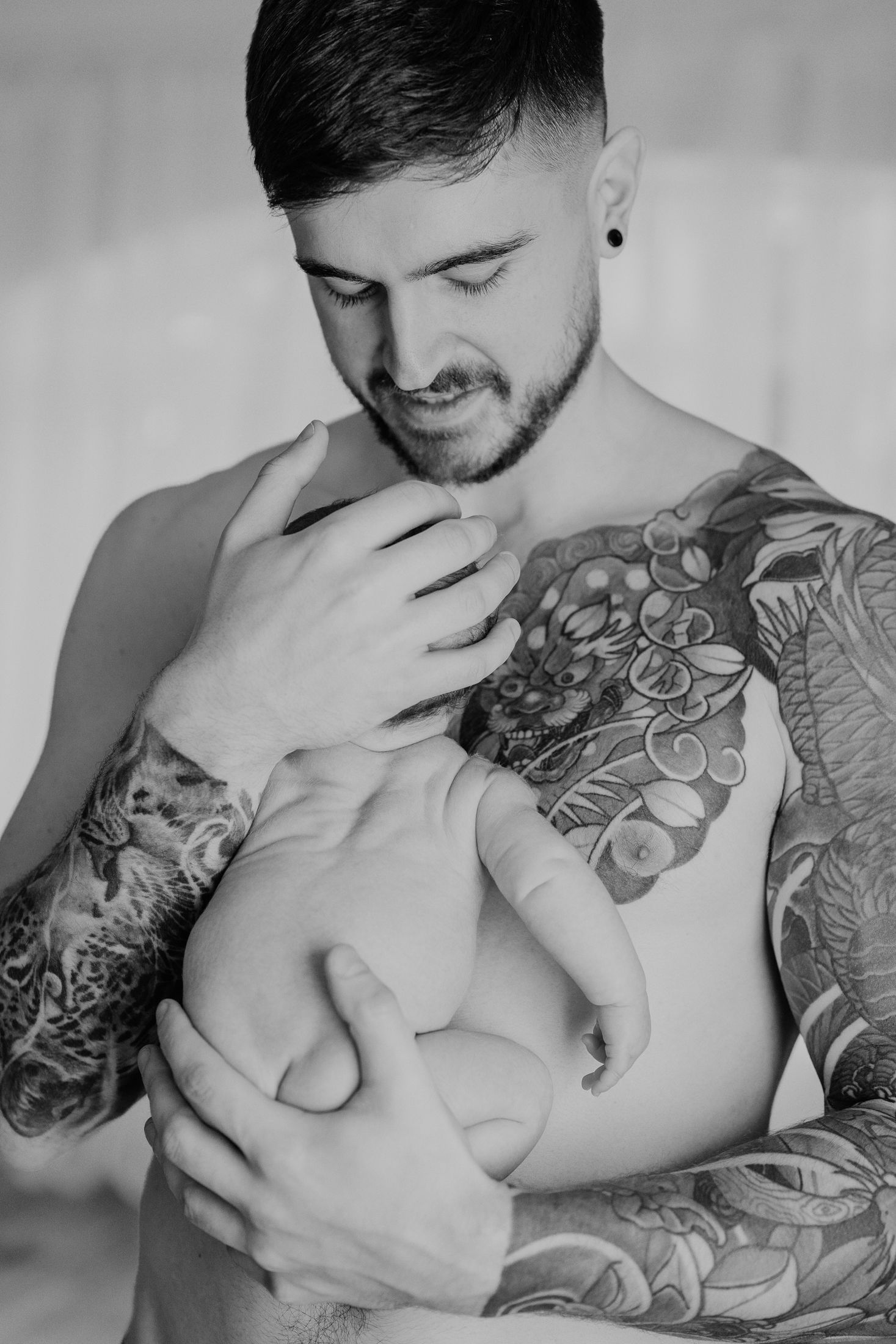 Black and white portrait of shirtless Dad with  tattoos holding his baby, natural newborn photography Dublin.