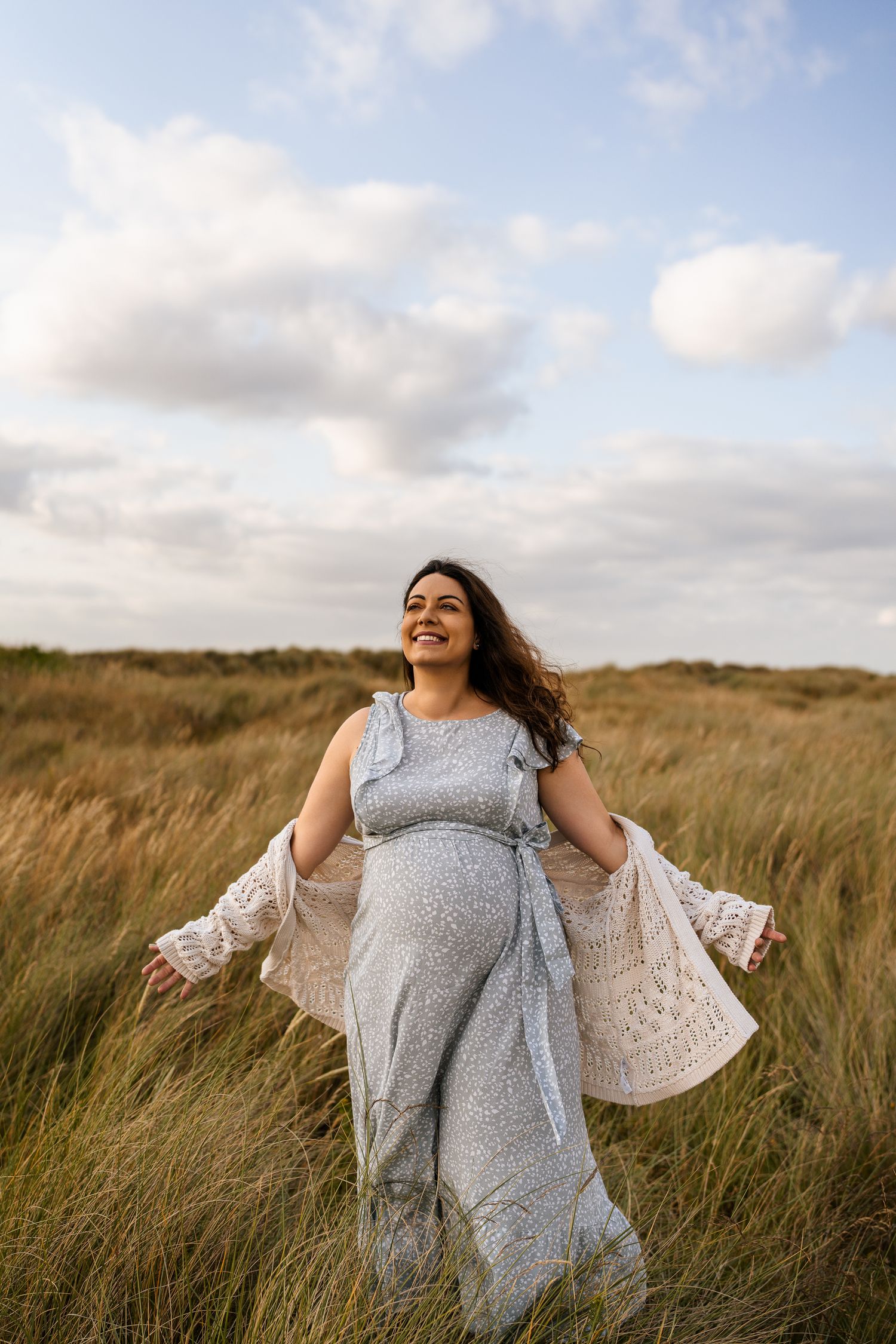 Woman in flowy dress twirls joyfully in tall grass field under cloudy sky.