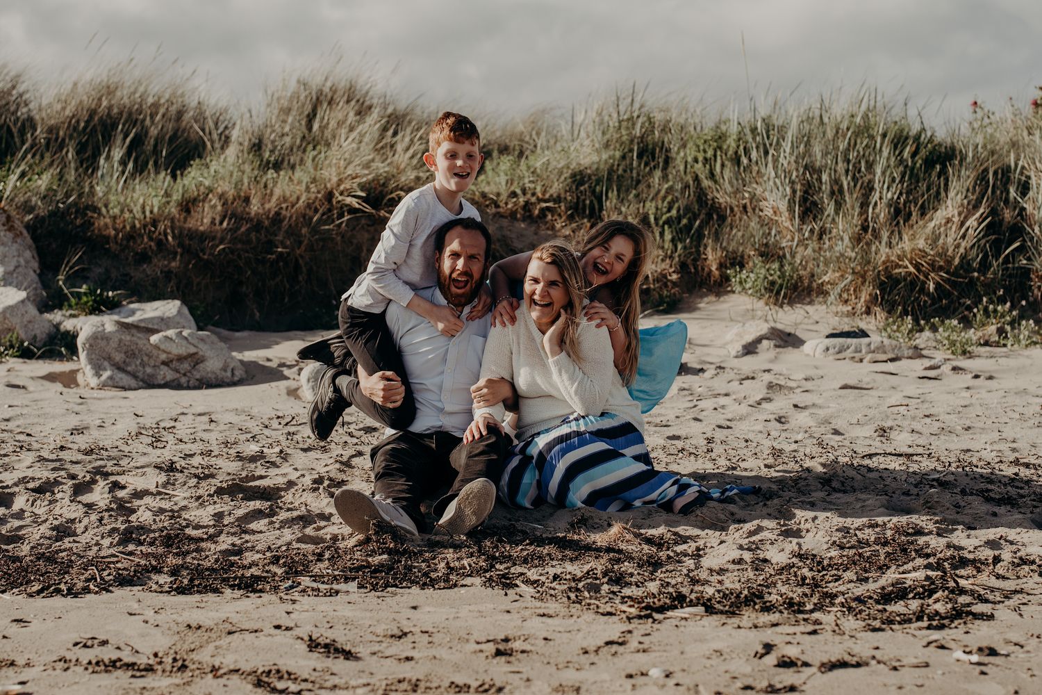 Family relaxing and playing together on a sandy beach near grass-covered dunes.