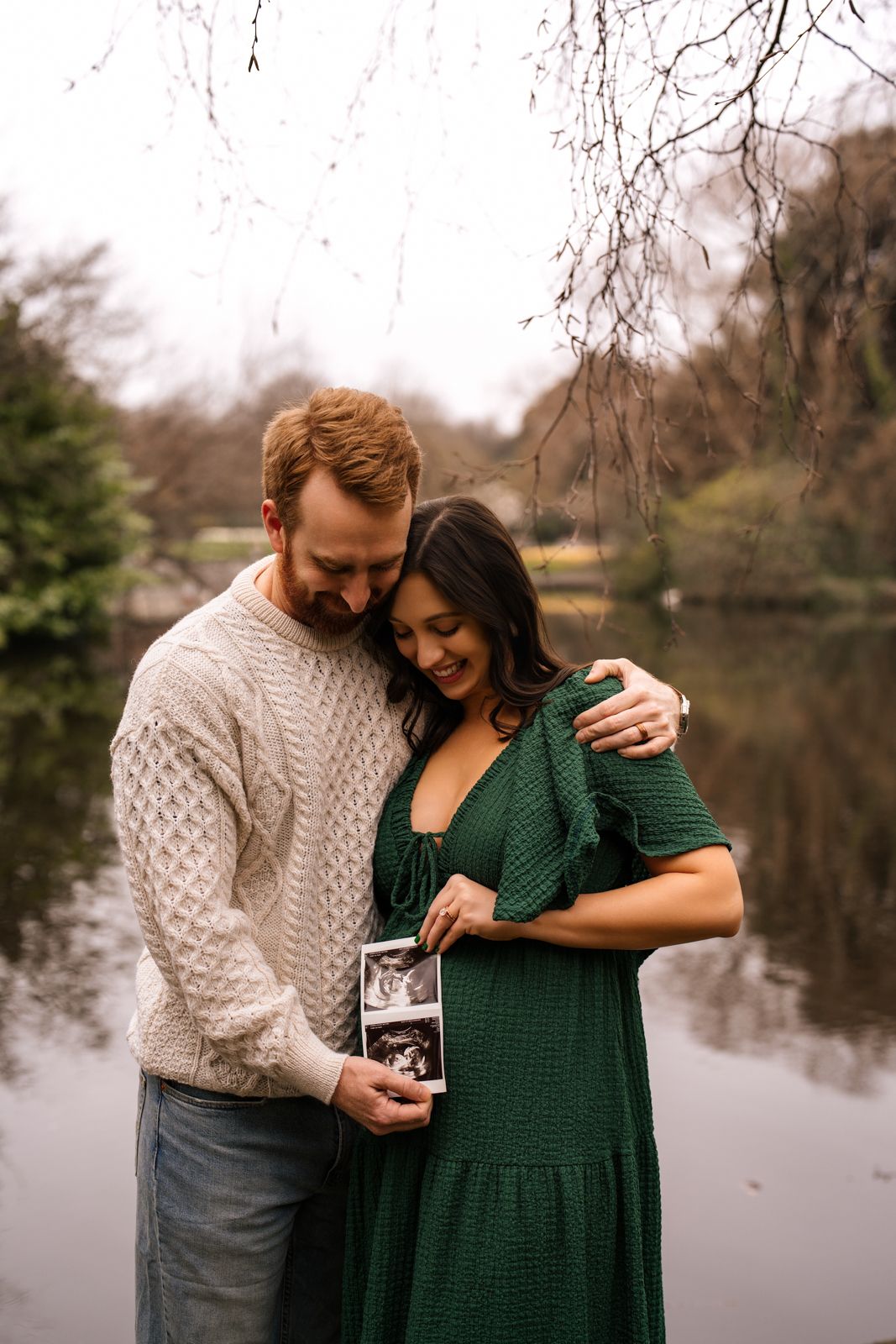 Couple holding ultrasound photo by a lake, woman in green dress, man in cream sweater, announcing pregnancy outdoors.