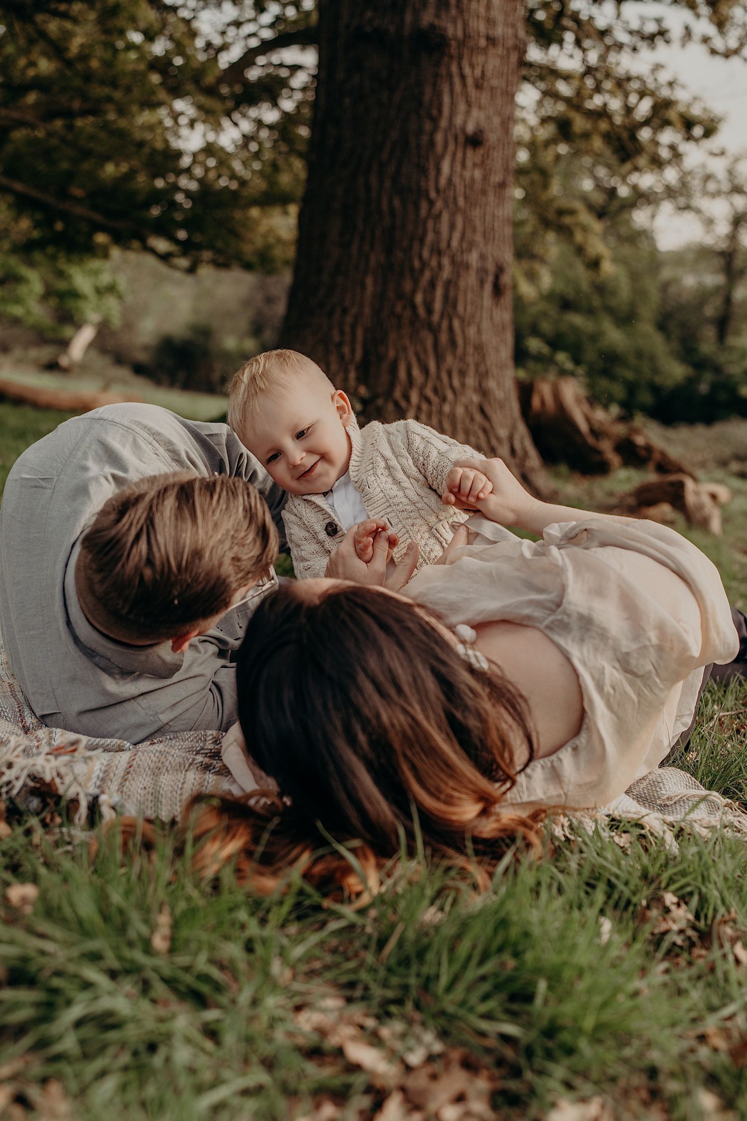 Family lying together on grass under a large tree in a park.