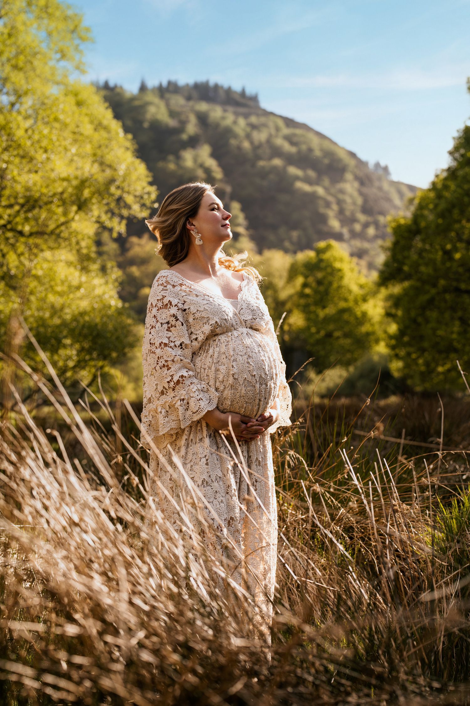 Person in lace shawl poses in golden sunlight among tall grasses with mountains in background.