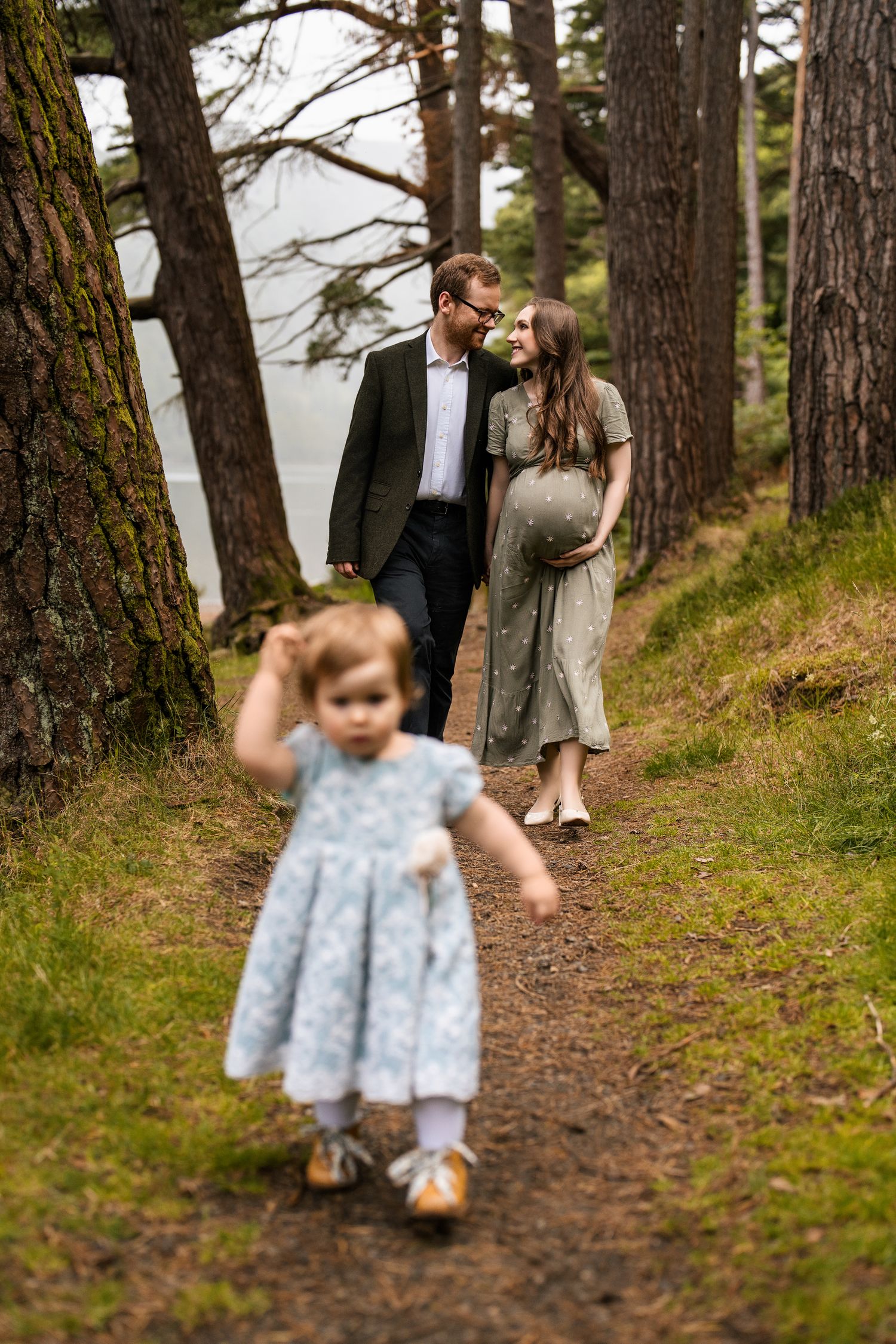 A small child runs along a forest path while adults watch from behind.