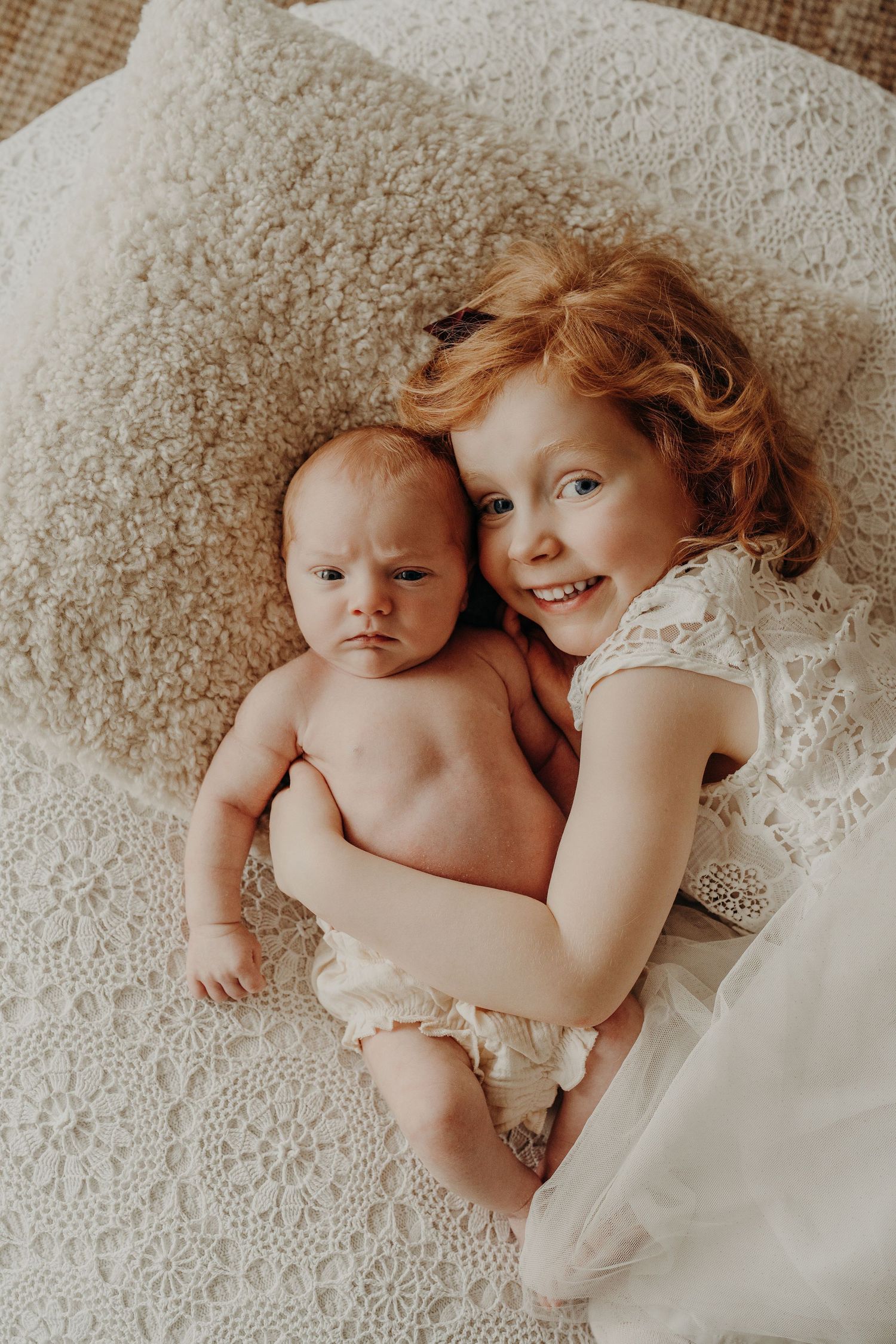 Intimate portrait of siblings lying together on a plush carpet in white clothing.
