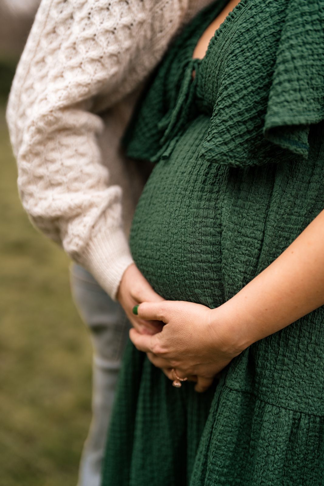 A pregnant woman wearing a green dress and a beige sweater, standing in a field.