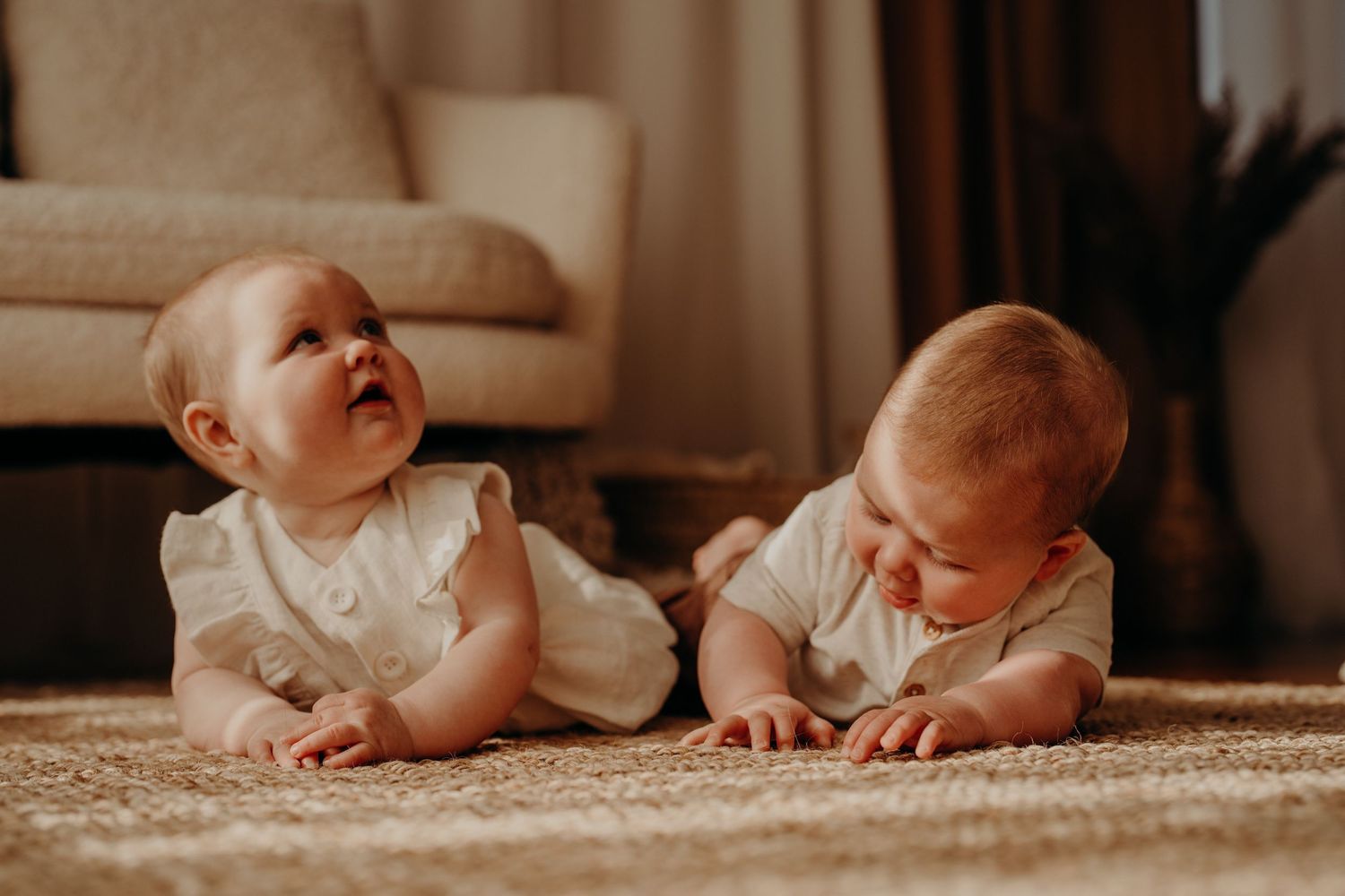Baby in white clothing crawls on beige carpet in sunlit room.