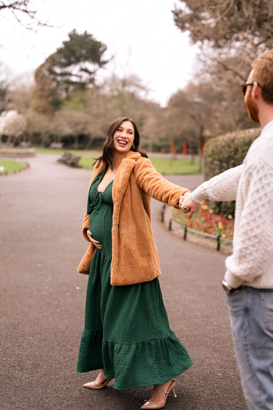 Pregnant woman in green dress and tan coat holds hands with partner in a park setting.