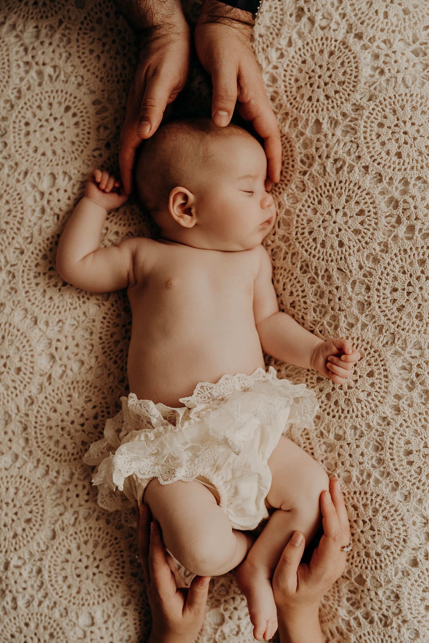 Newborn baby lying on lace blanket with caring hands providing gentle support.