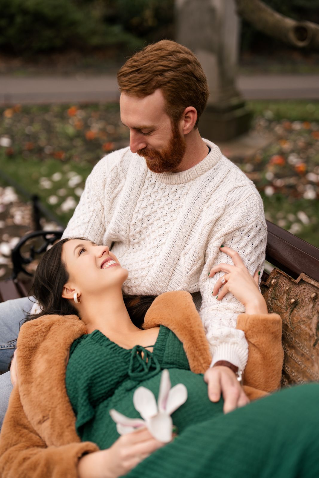 A couple relaxes on a park bench, woman in green dress lying back smiling, man in white cable-knit sweater gazing down at her.