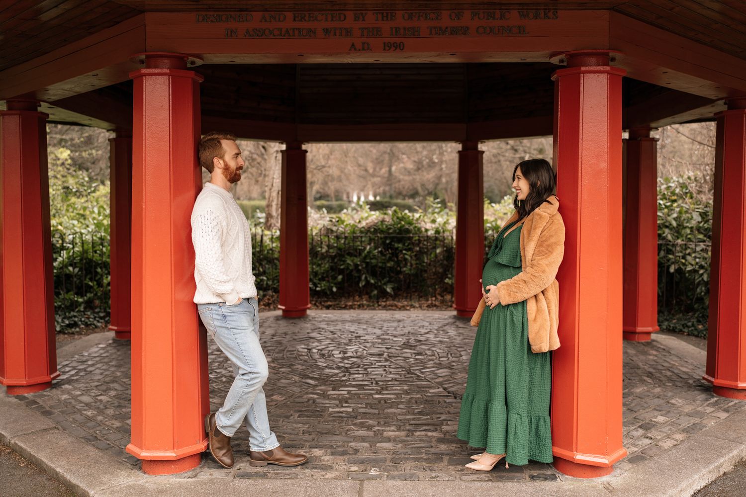 Two people stand under a red-pillared structure, conversing in an outdoor setting.