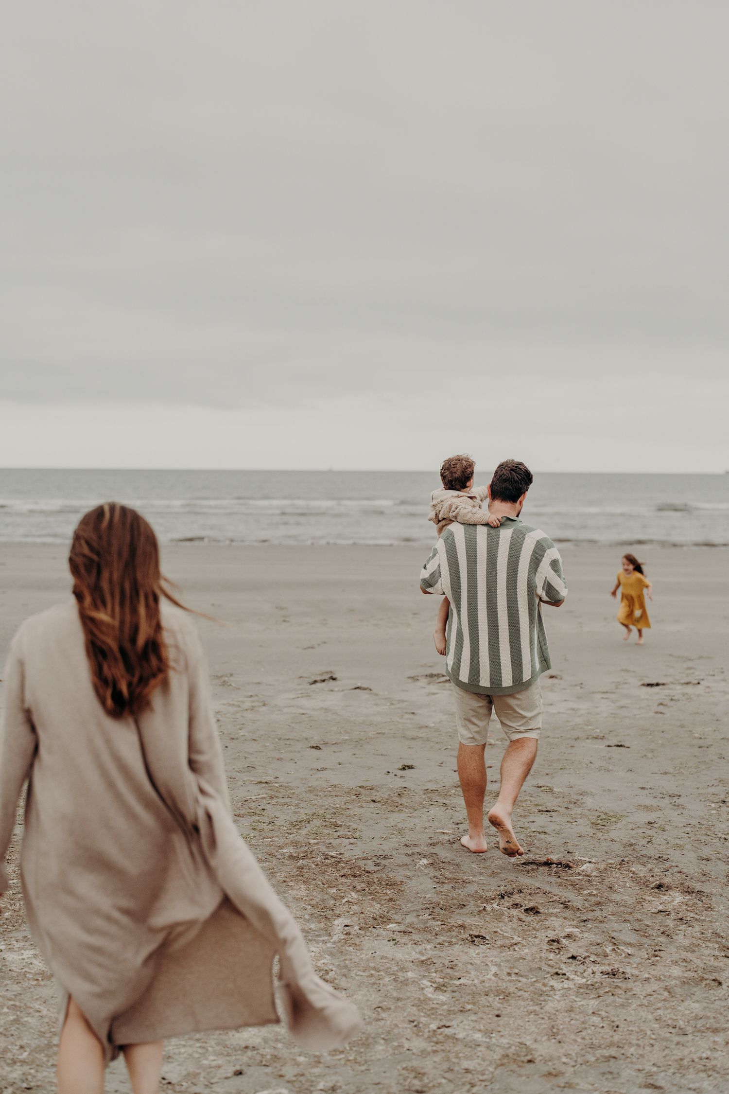 A family walks together on a misty beach holding hands and playing in casual beige and striped clothing.