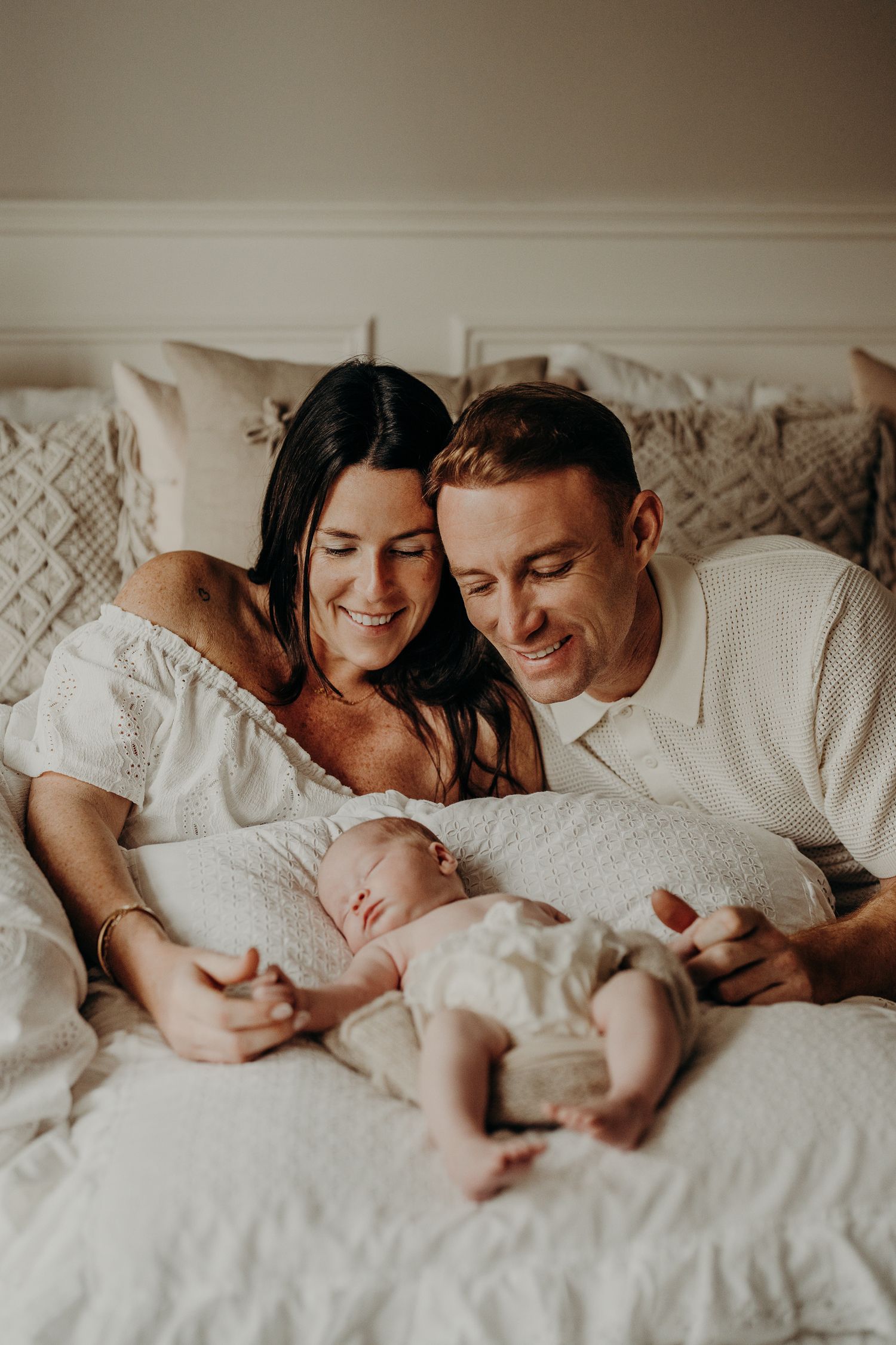 Family shares a joyful moment together on white bedding with their sleeping newborn.