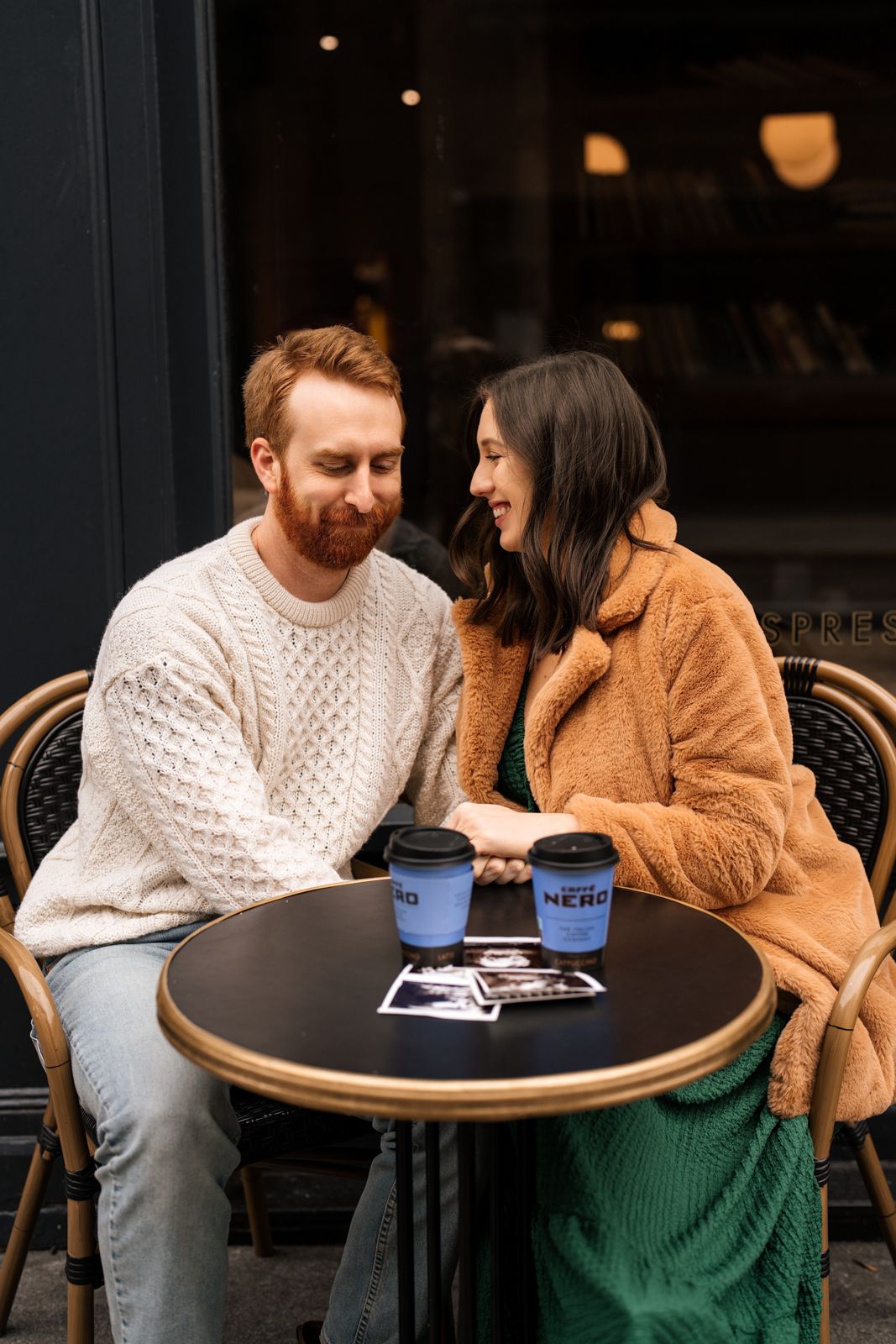 Couple sitting at outdoor cafe table with Caffe Nero cups and photos, man in white sweater, woman in tan teddy coat.