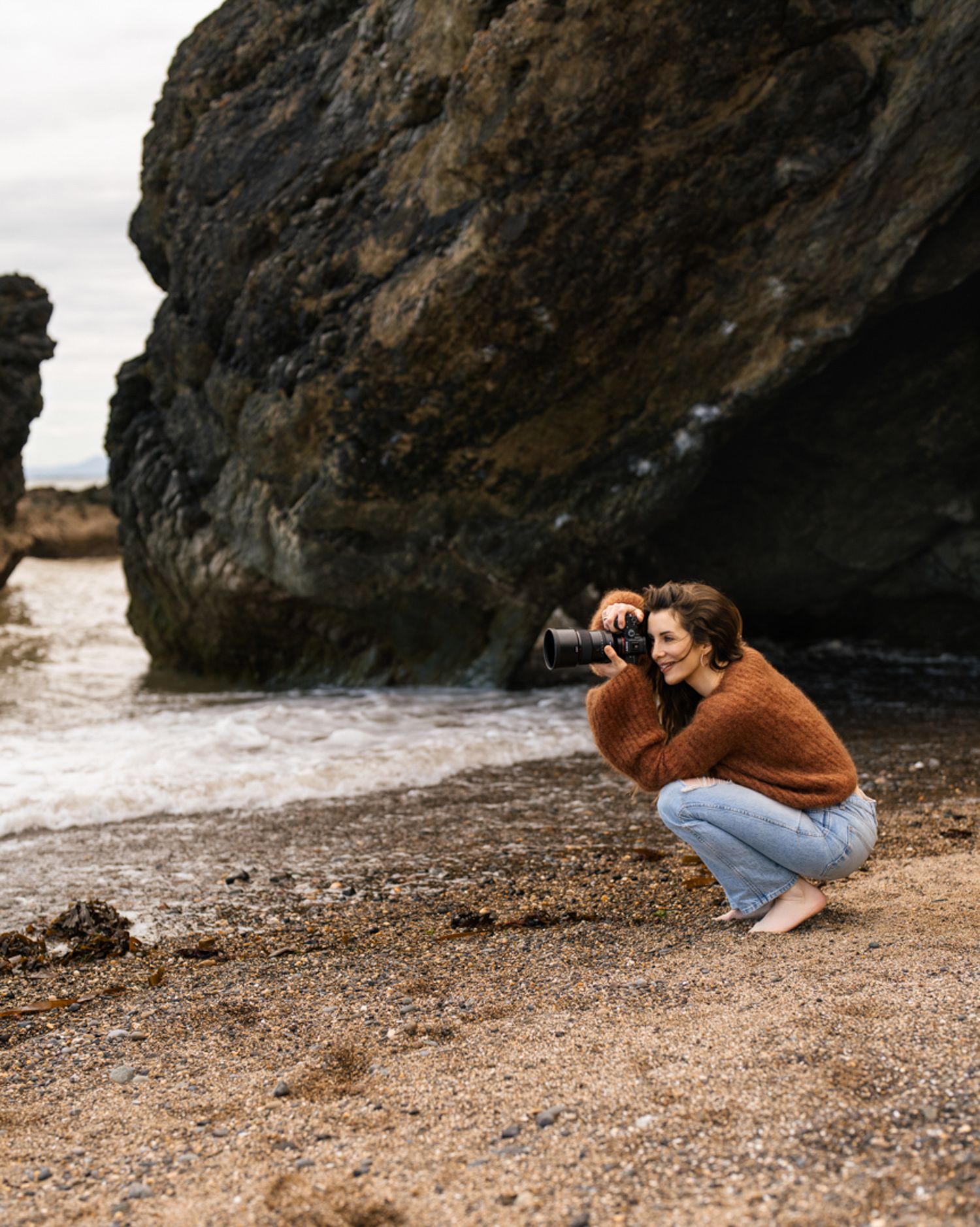 Person in brown sweater crouching to take photo by rocky coastal cliffs.