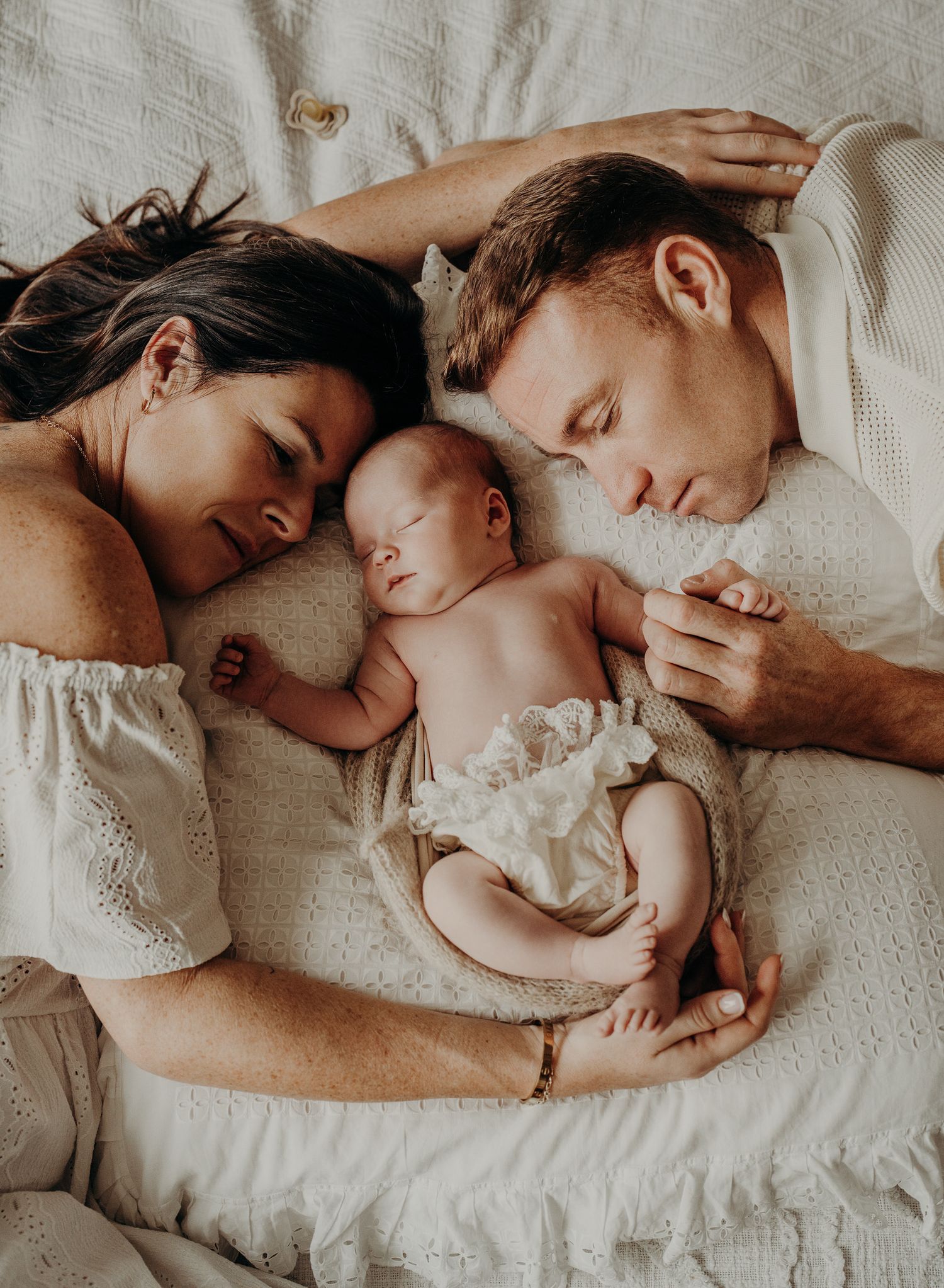 Intimate family moment as parents rest with their newborn baby on white bedding.