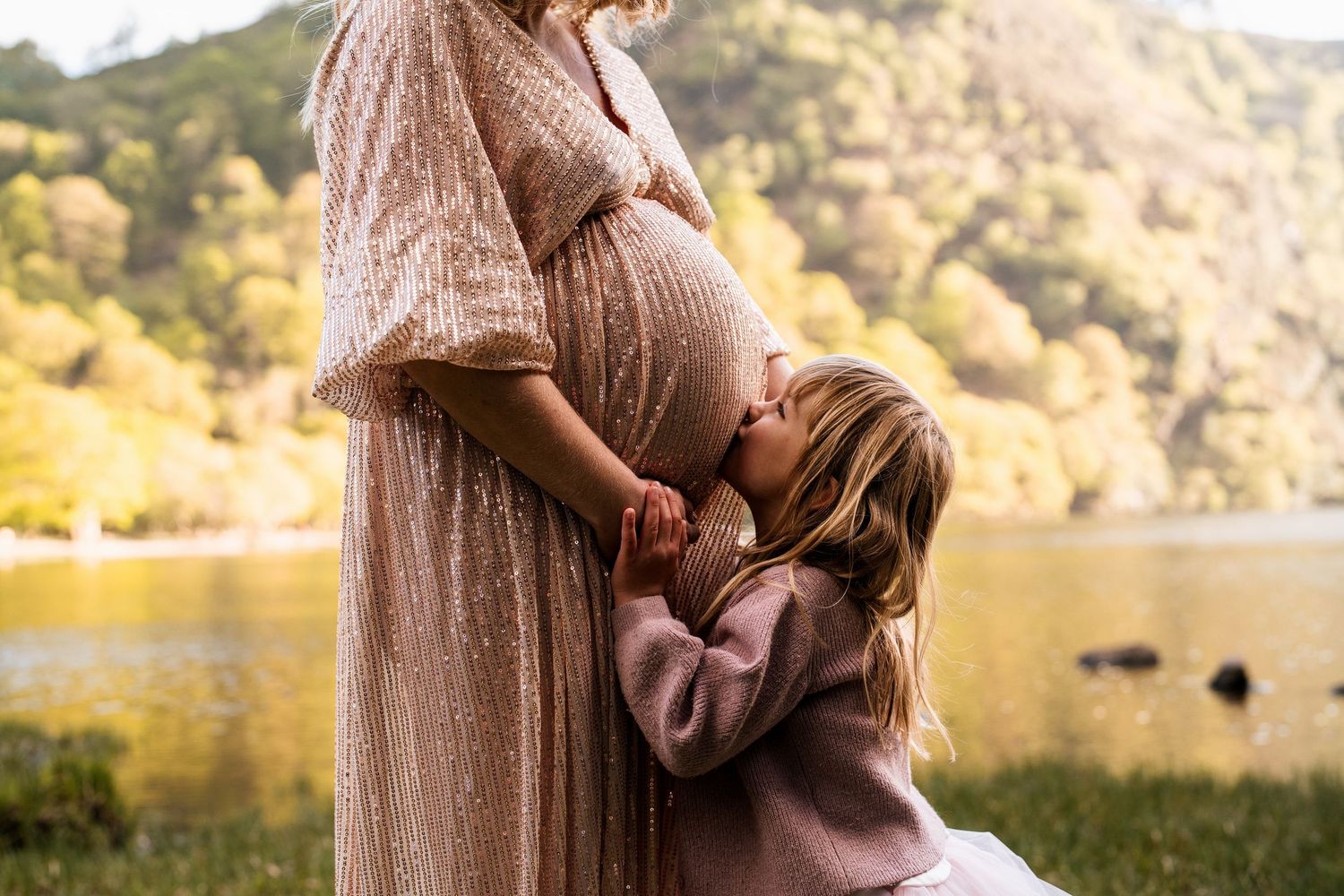 Maternity photos captured by a lake with mountains in the background during golden hour.