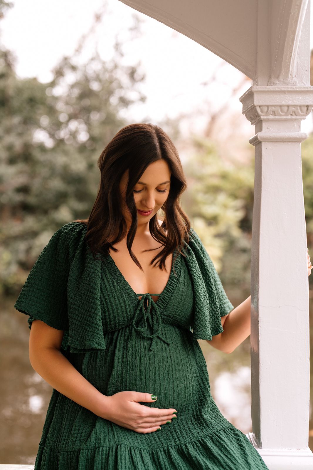 Pregnant woman in green dress cradling baby bump on porch.