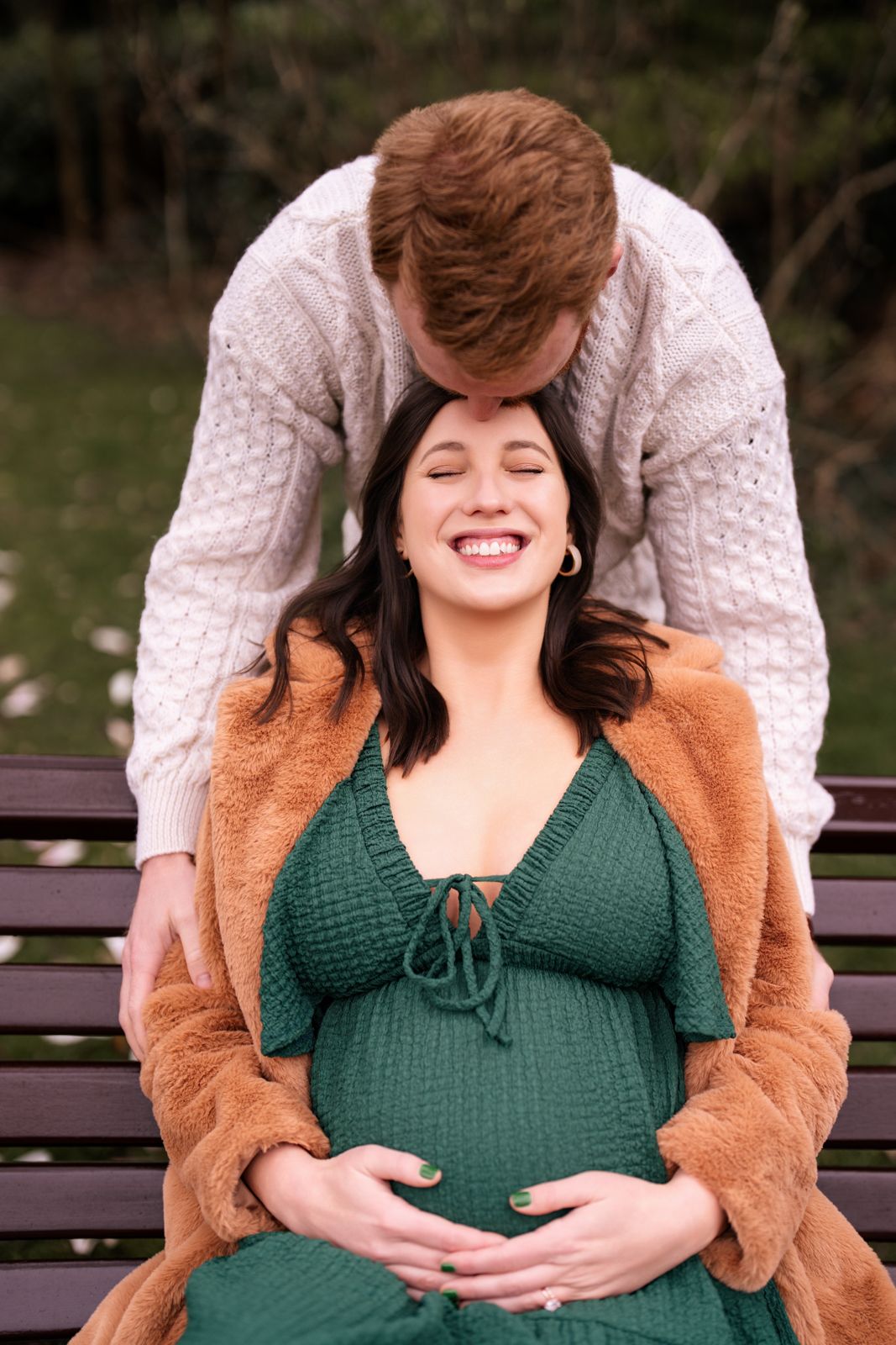 A pregnant woman in a green dress is embraced by a man wearing a gray sweater on a park bench.