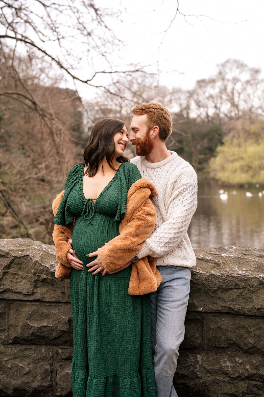 A pregnant woman and her husband embrace affectionately in a park setting with trees and water in the background.