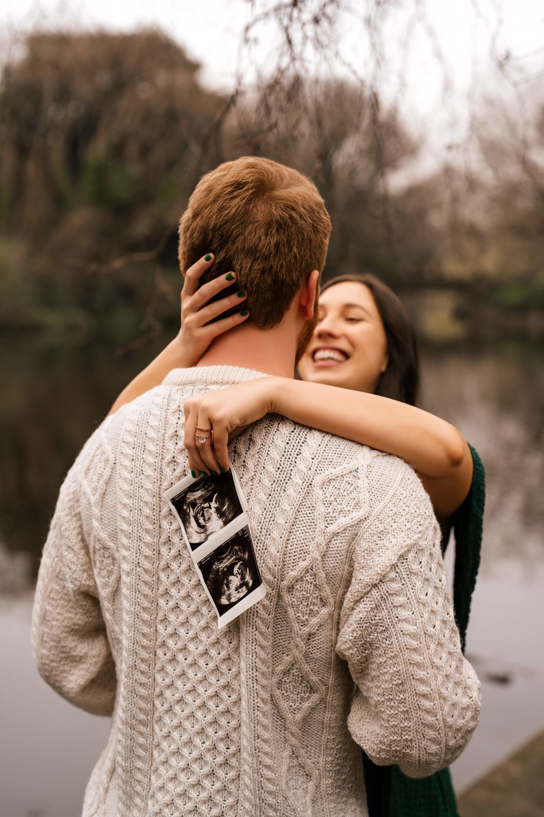 Happy couple embracing outdoors holding ultrasound photos, pregnancy announcement near a lake in autumn.
