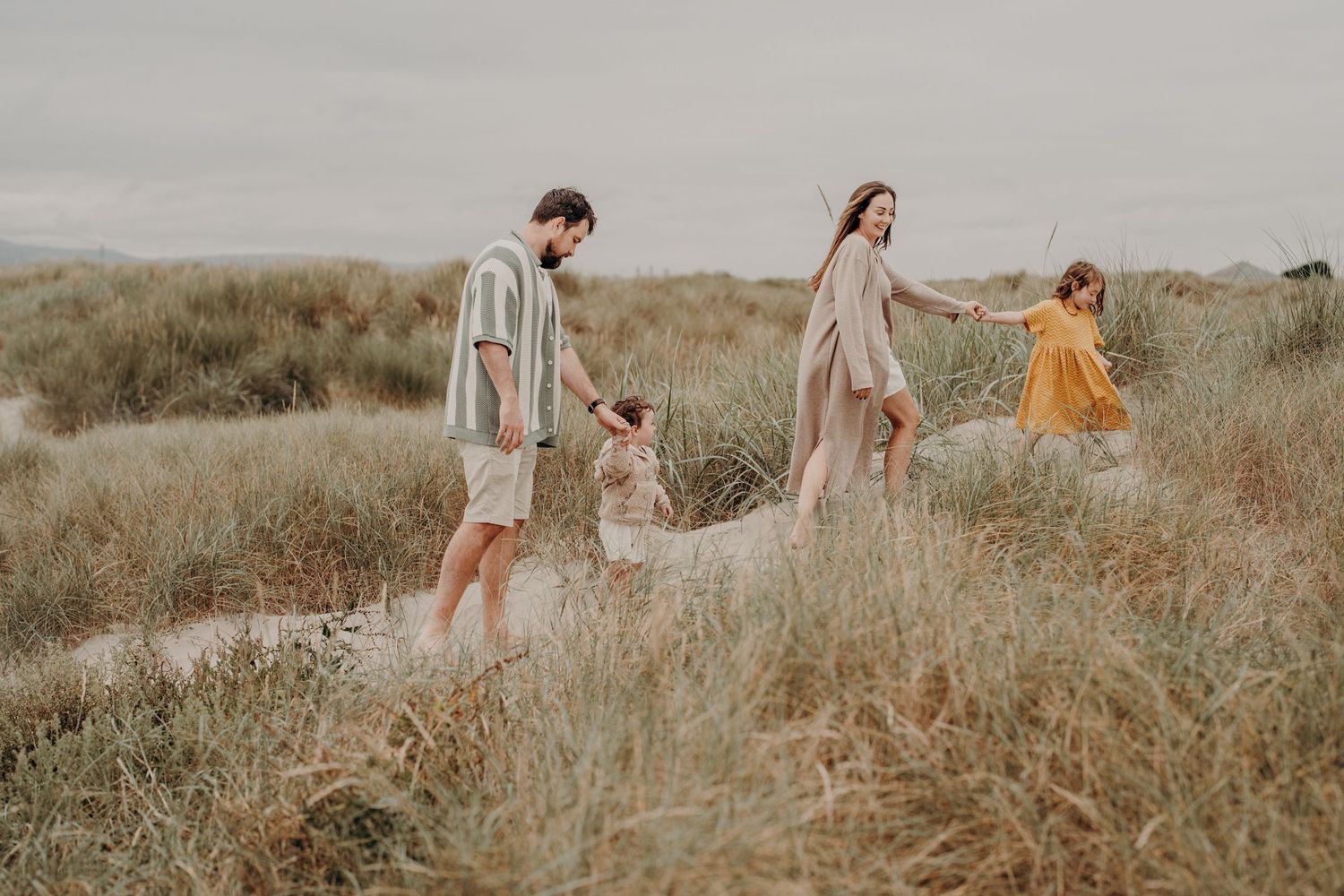 Family walking through coastal dunes on an overcast day wearing earth-toned clothing.