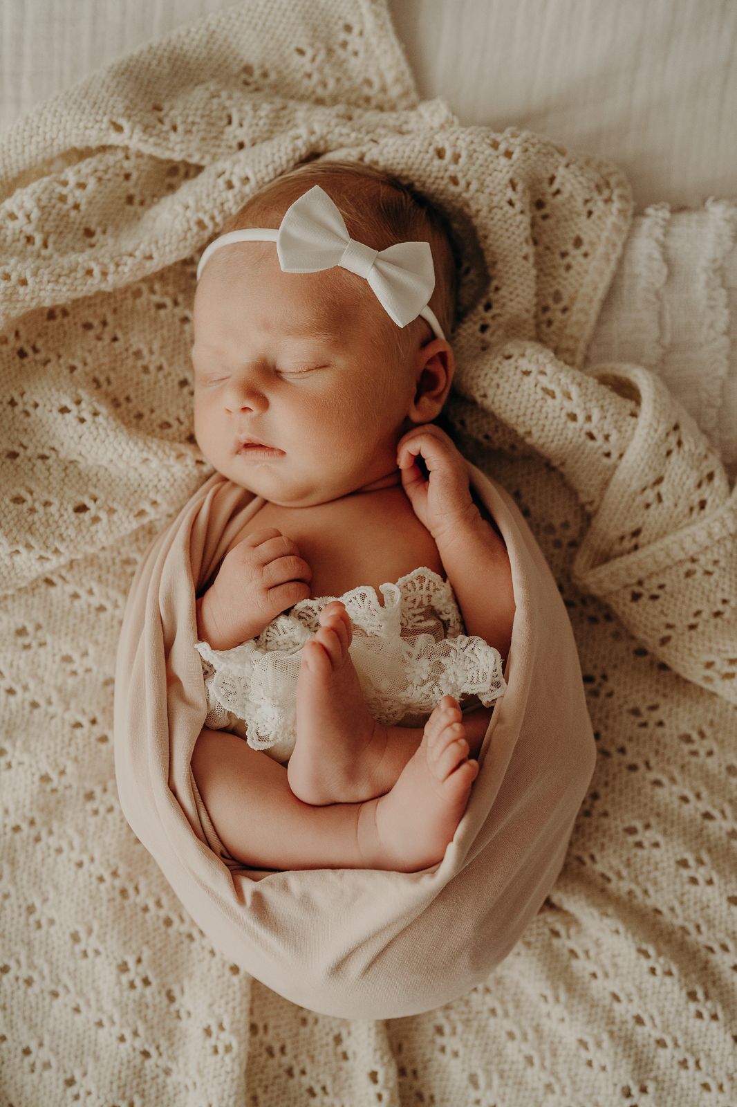 Newborn baby wearing white bow headband wrapped in beige fabric on lace blanket.