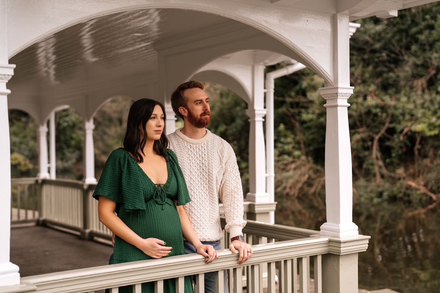Pregnant couple in green dress and cream sweater posing on white gazebo bridge surrounded by lush greenery.