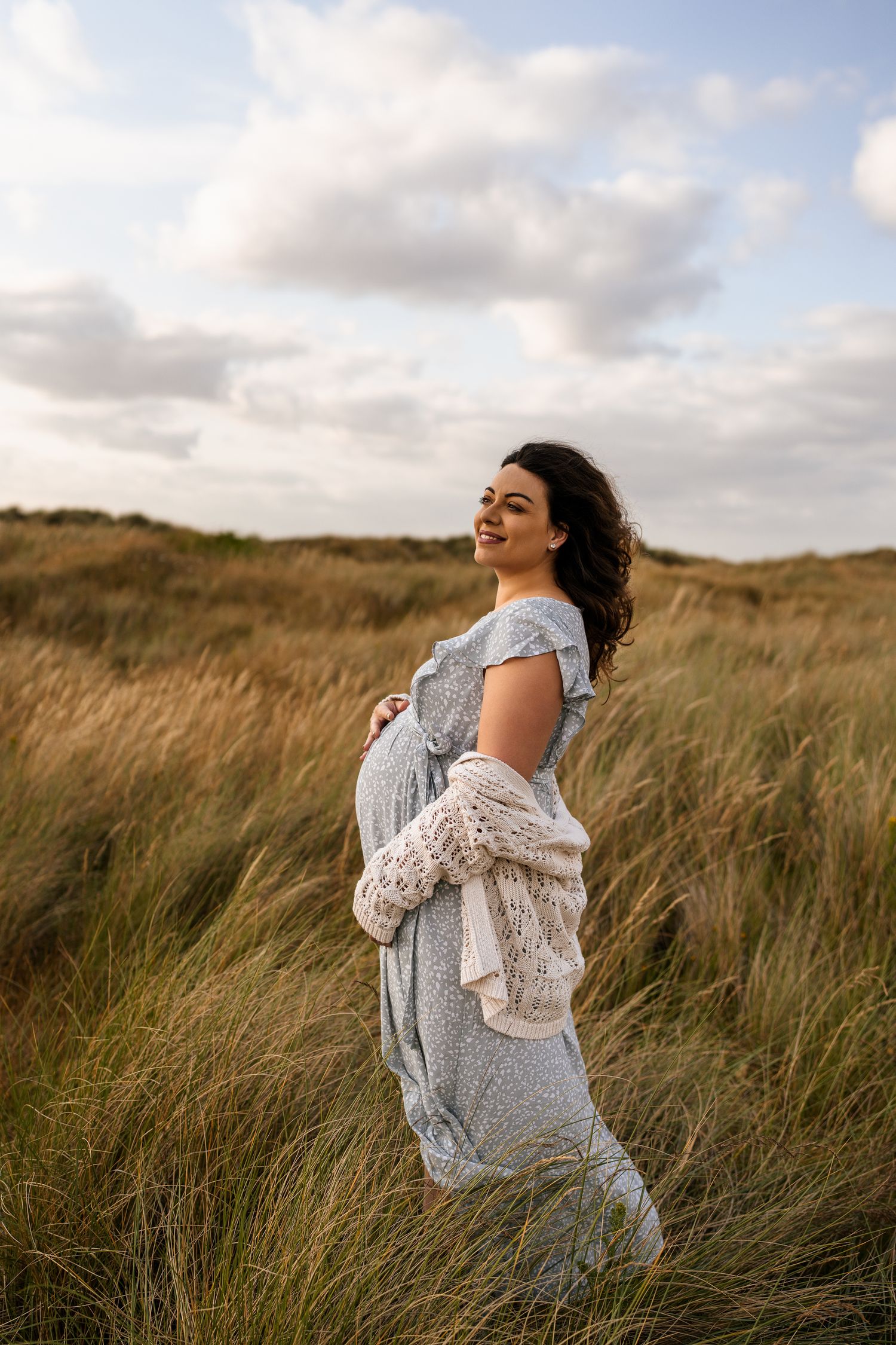 A pregnant woman in a gray dress poses in a windswept field at sunset.