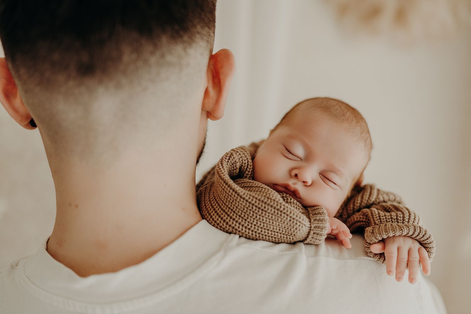A sleeping baby in a brown knit sweater rests peacefully on Daddy's shoulder in soft natural light.
