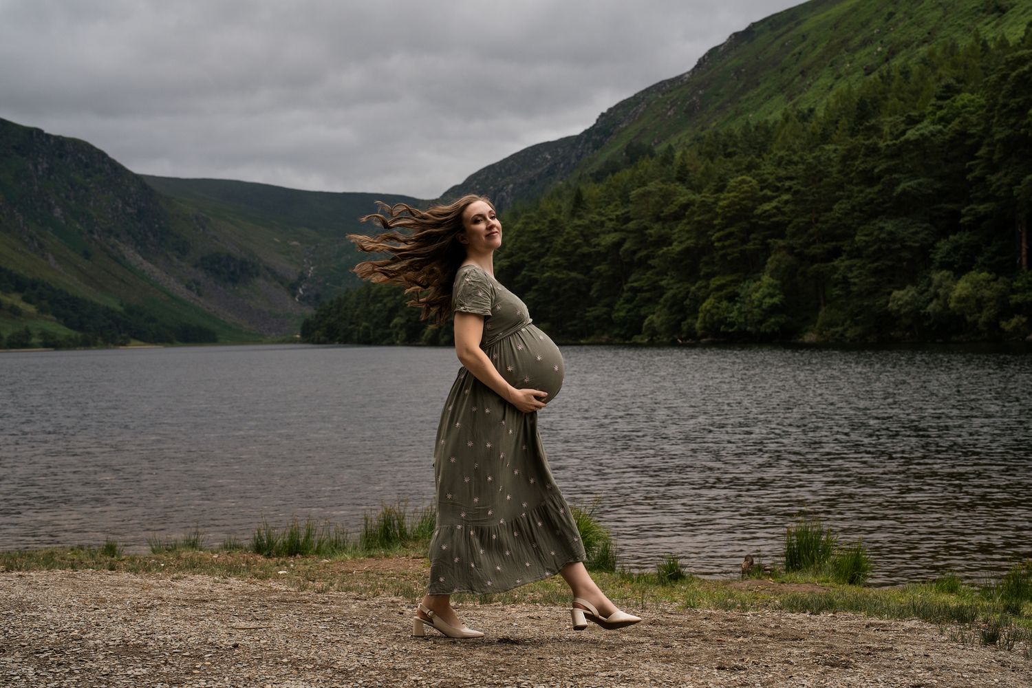 Expectant woman in flowing dress stands by a misty mountain lake with wind in her hair.