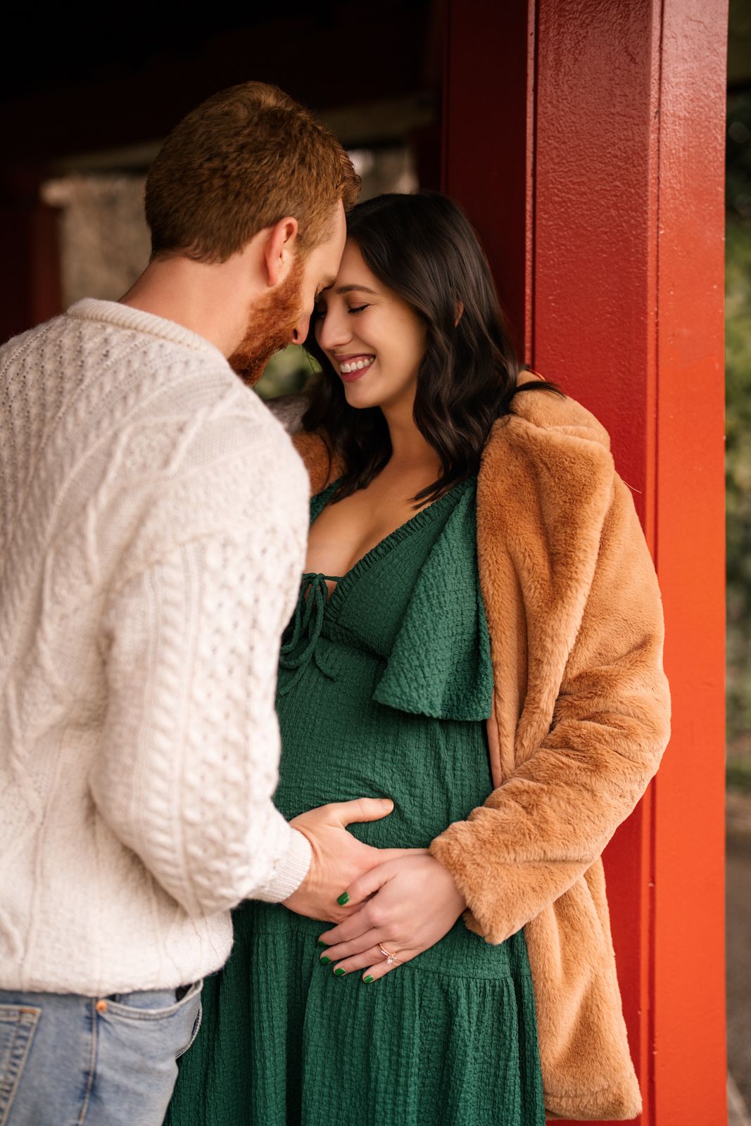 A smiling pregnant woman in a green dress and tan coat leans against a red wall as her partner tenderly holds her baby bump.