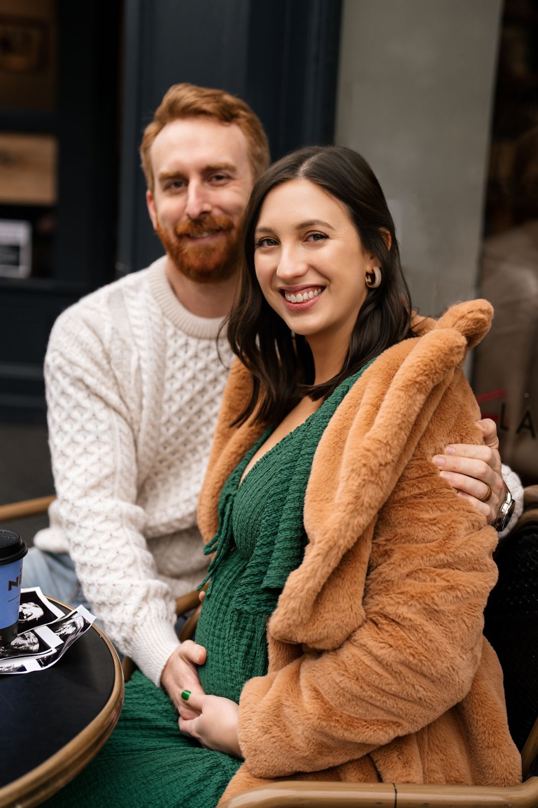 Smiling pregnant woman in green dress and fur coat sits with redheaded bearded man in white sweater at outdoor cafe.