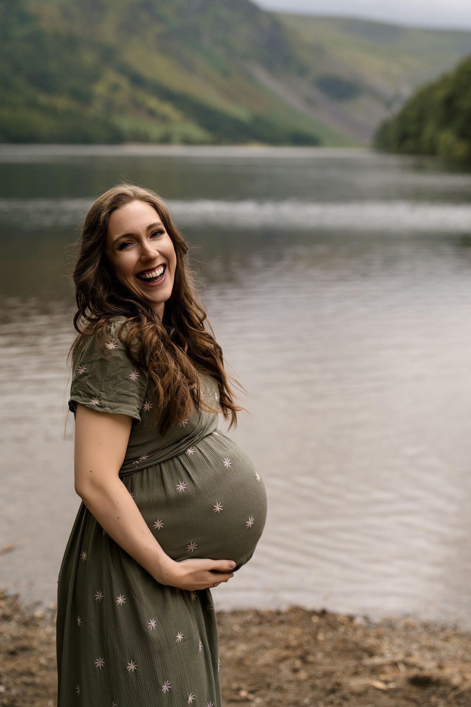 A pregnant woman in a green dress poses by a scenic mountain lake.