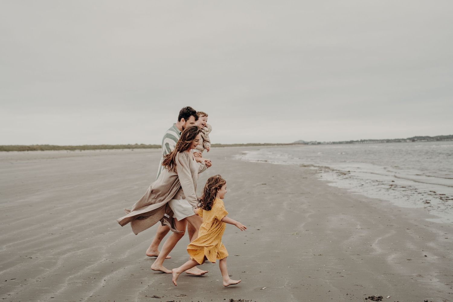 Family members run and play together along the shoreline on a cloudy beach day.