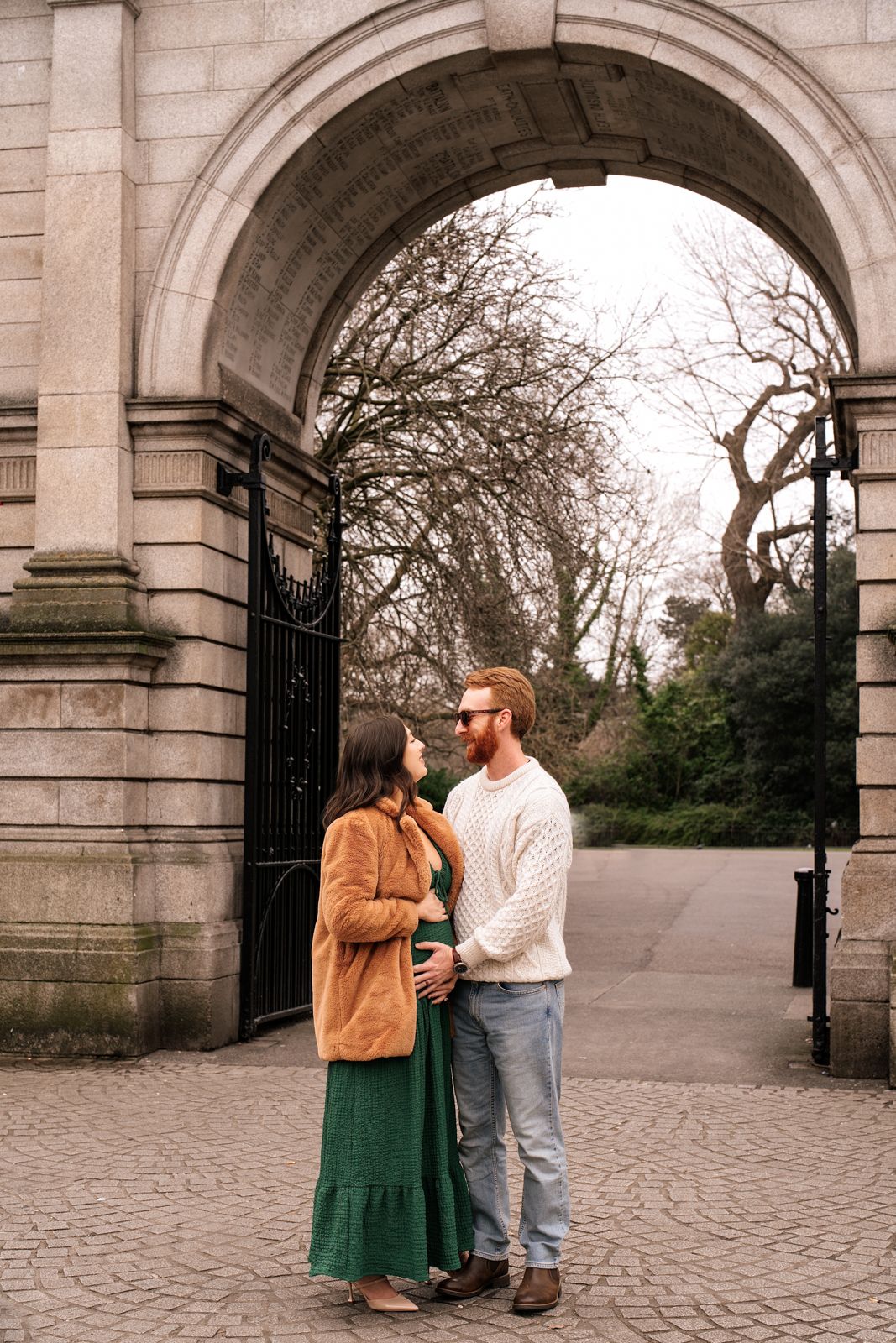A young couple stands in front of an ornate stone archway in a park, surrounded by bare trees on a cloudy day.