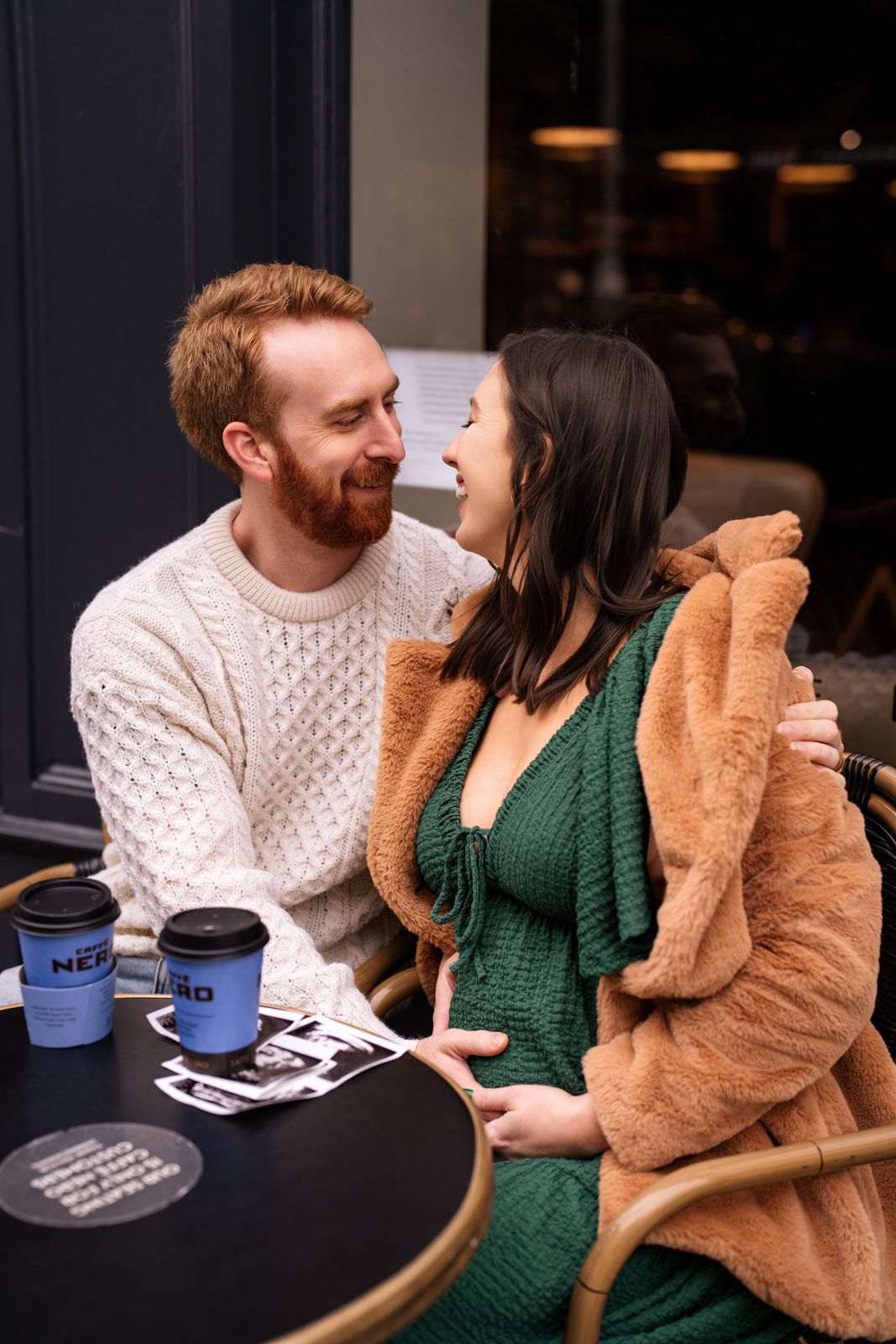 Happy couple sharing a joyful moment at a Caffe Nero outdoor table, woman in green dress and fur coat touching baby bump.