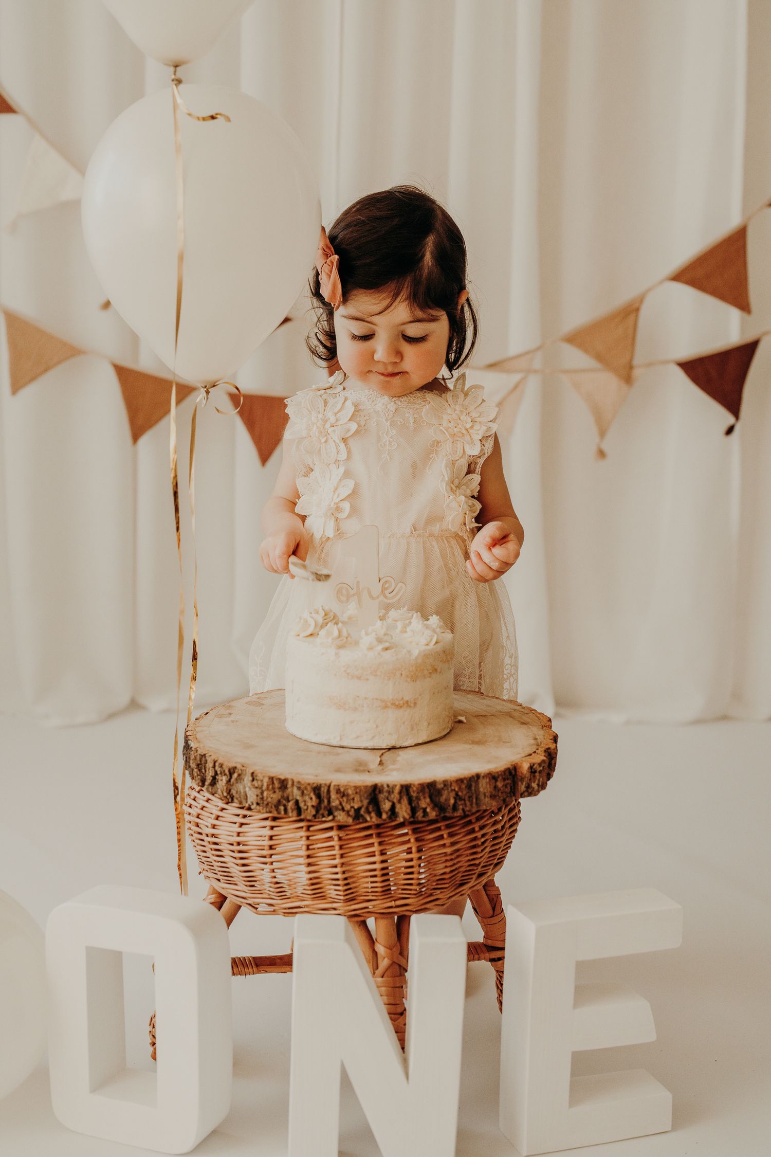 White birthday cake with delicate lace details sits in a woven basket against a light backdrop with hanging decorations.