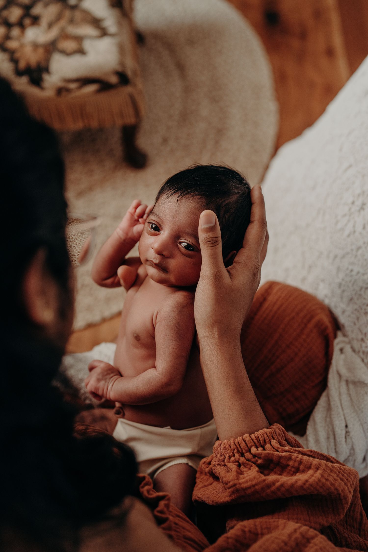 Gentle hands holding and comforting a newborn wrapped in rust-colored fabric.