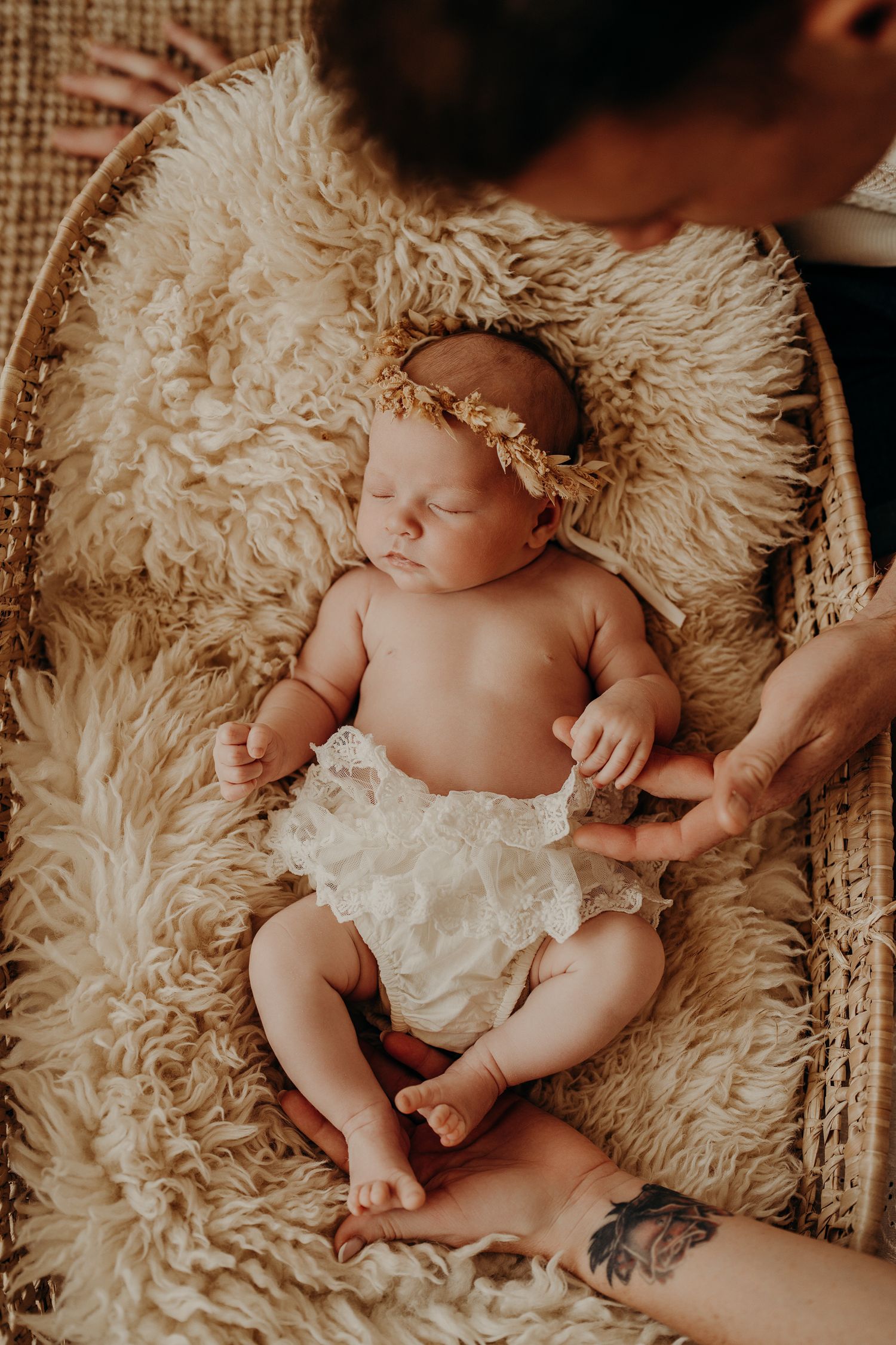 Sleeping newborn wearing a floral crown and white bloomers rests on fluffy cream-colored fabric.