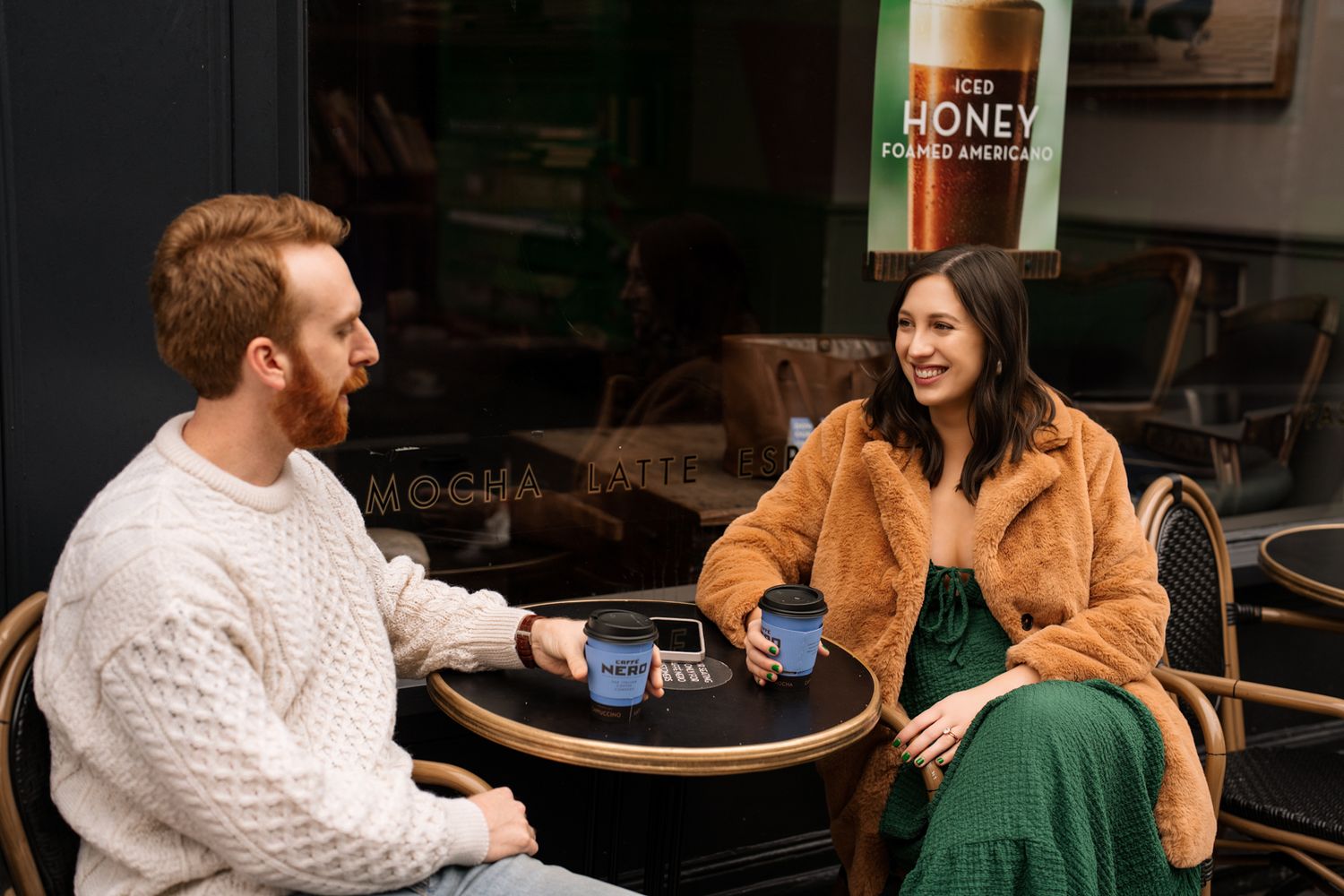 A man and woman enjoy coffee at an outdoor cafe table, smiling and chatting in stylish casual outfits.