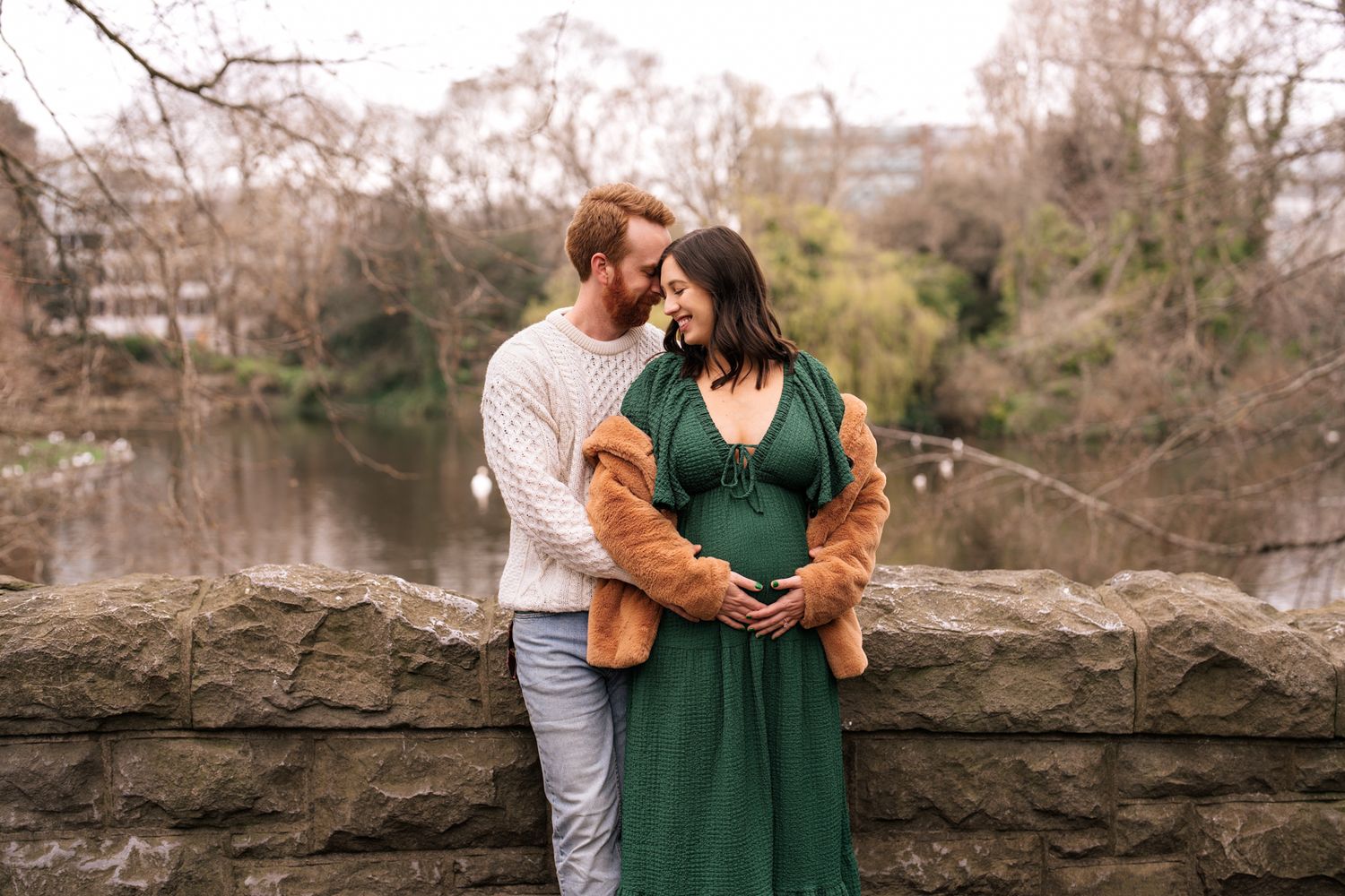 A couple embracing each other affectionately near a body of water with trees in the background.