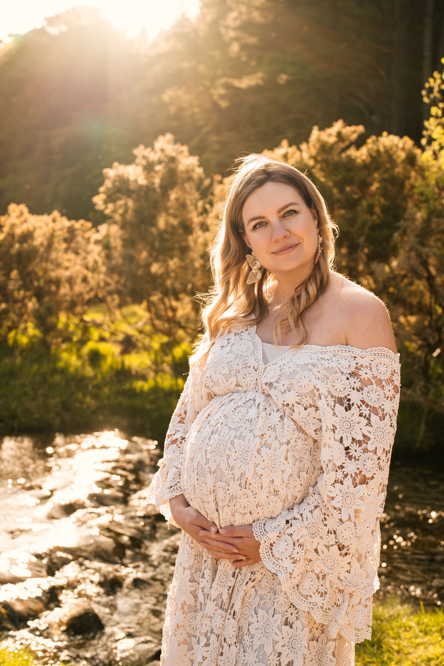 A golden sunlit maternity photoshoot featuring a white lace off-shoulder dress against a natural outdoor backdrop.