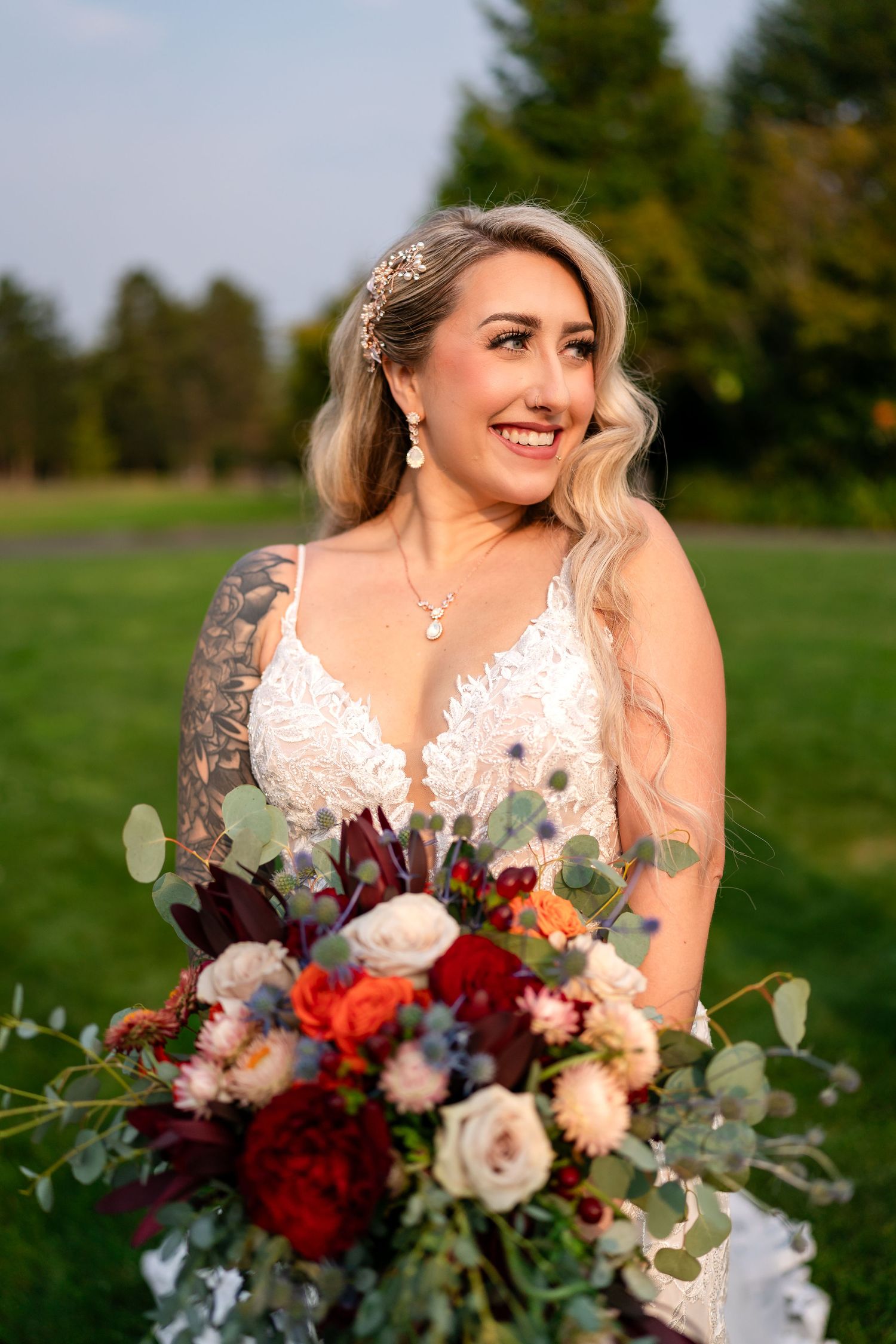 A bride in a white lace dress holds a stunning burgundy and blush bouquet with eucalyptus in an outdoor setting.