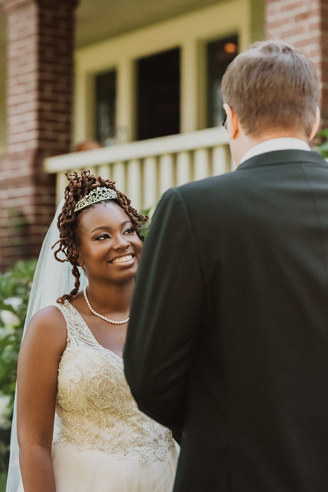 A bride in a lace wedding dress and pearl necklace smiles joyfully during an outdoor wedding celebration.