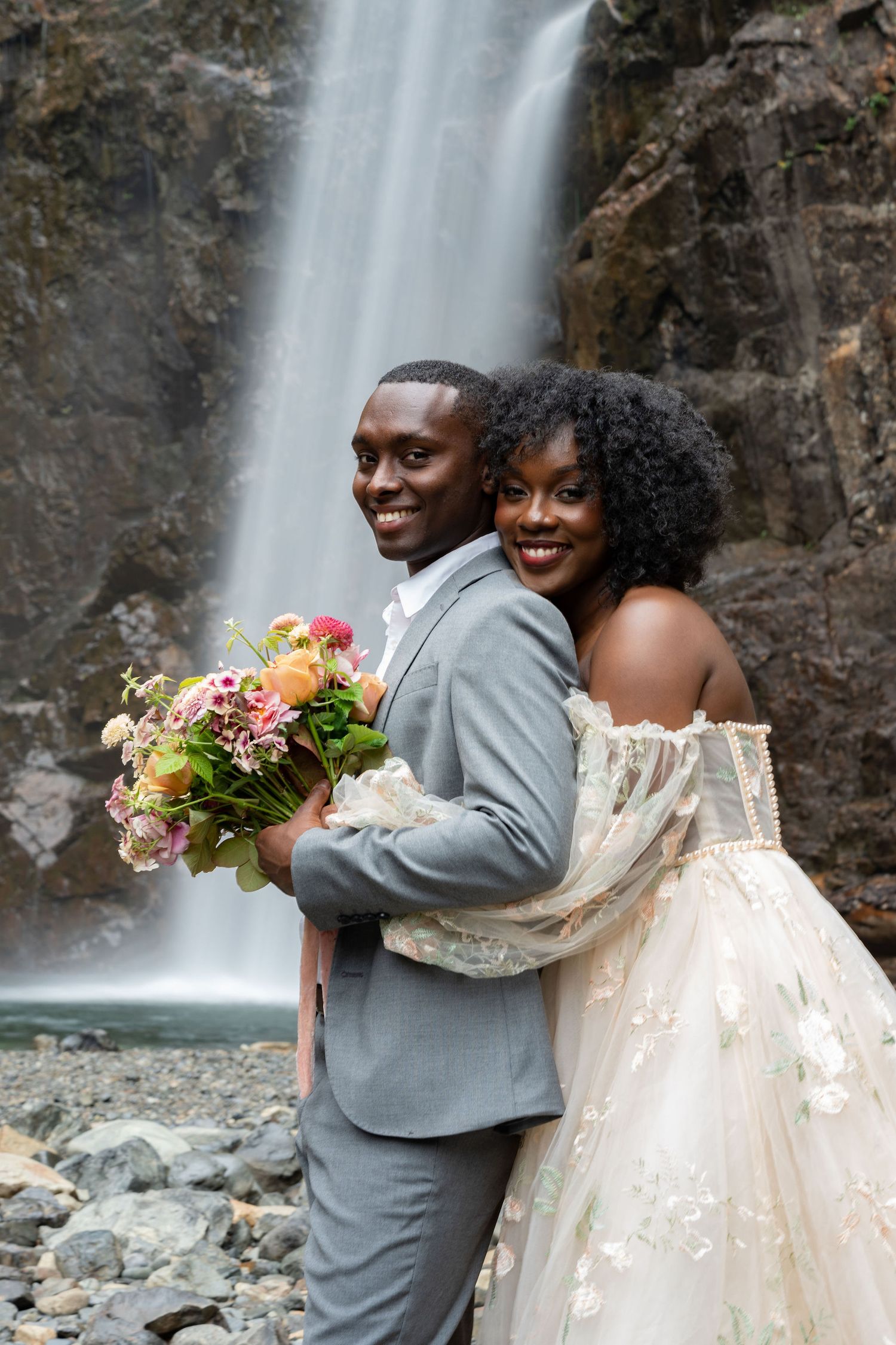 A joyful wedding couple embraces in front of a cascading waterfall.