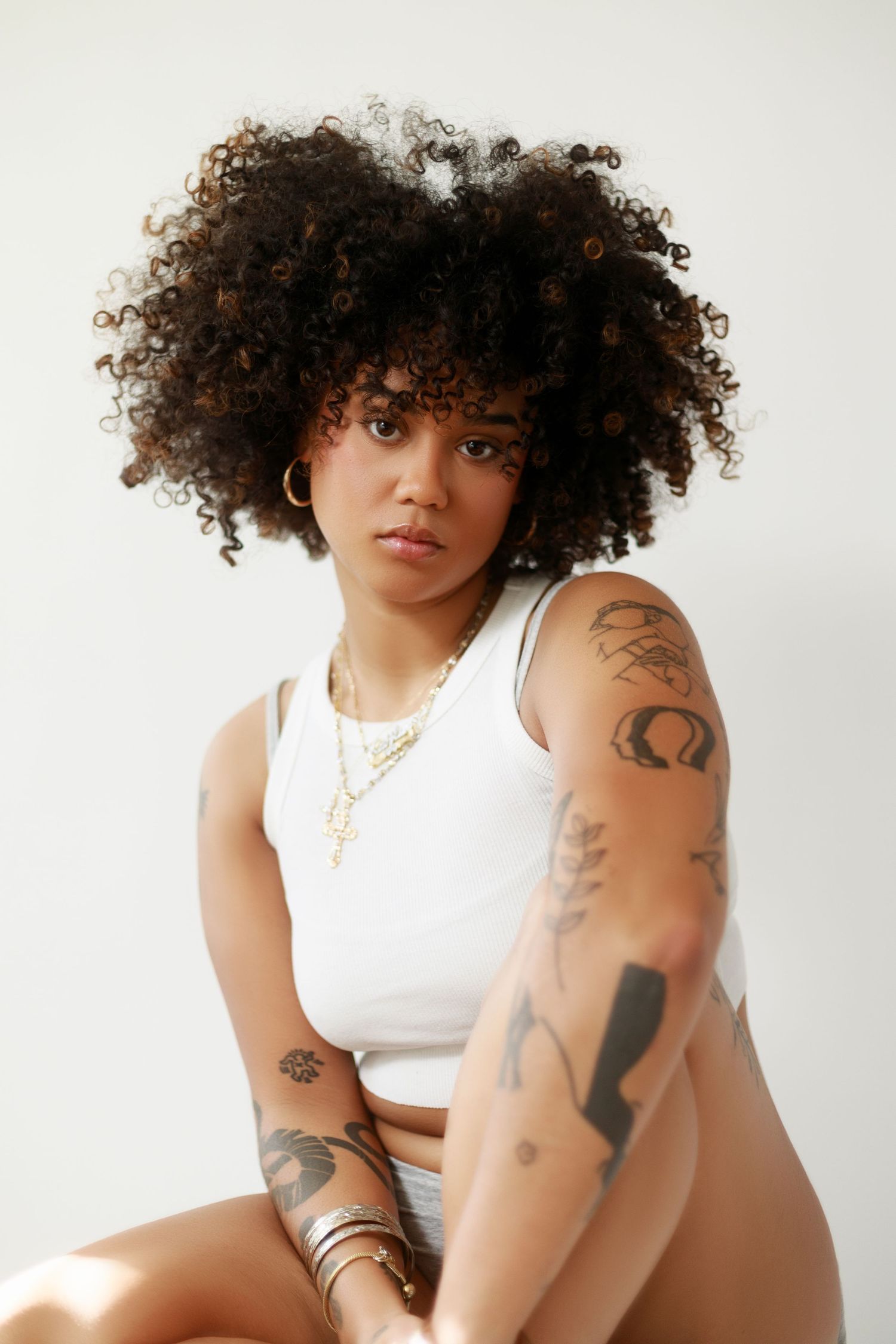 Model with natural curls and arm tattoos poses confidently in white crop top.