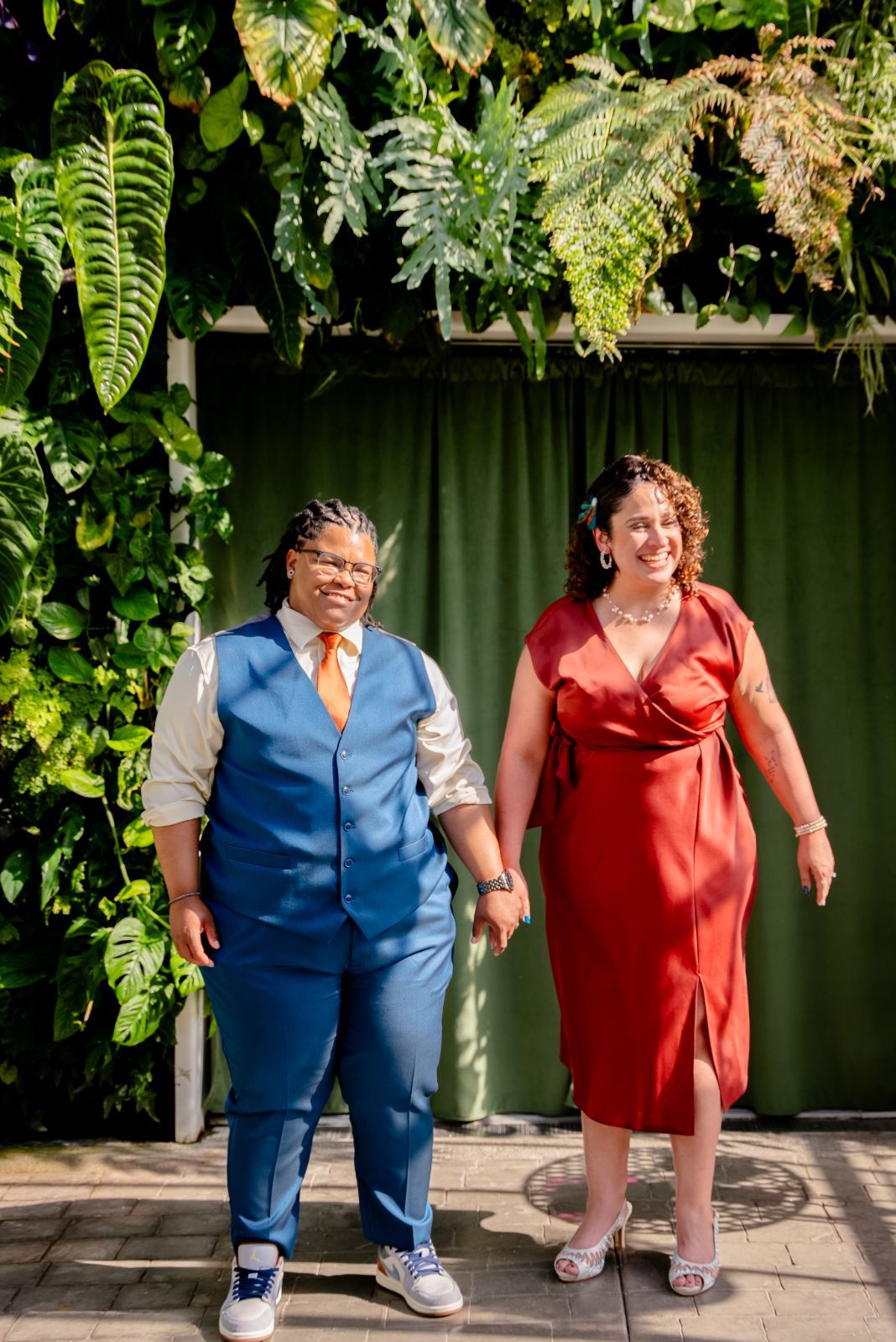 People in formal attire stand against a dark wall with greenery overhead in a garden setting.