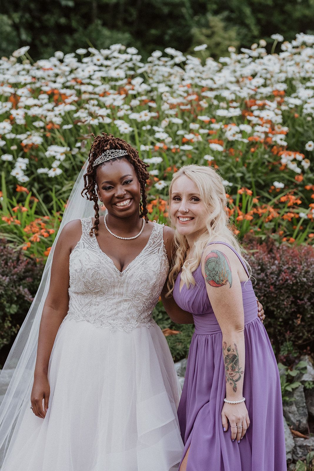 Wedding party poses in white and purple dresses among blooming daisies and orange flowers.