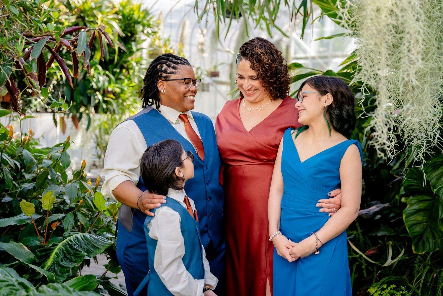 Three people in blue and red outfits pose together in a lush garden setting with tropical plants.